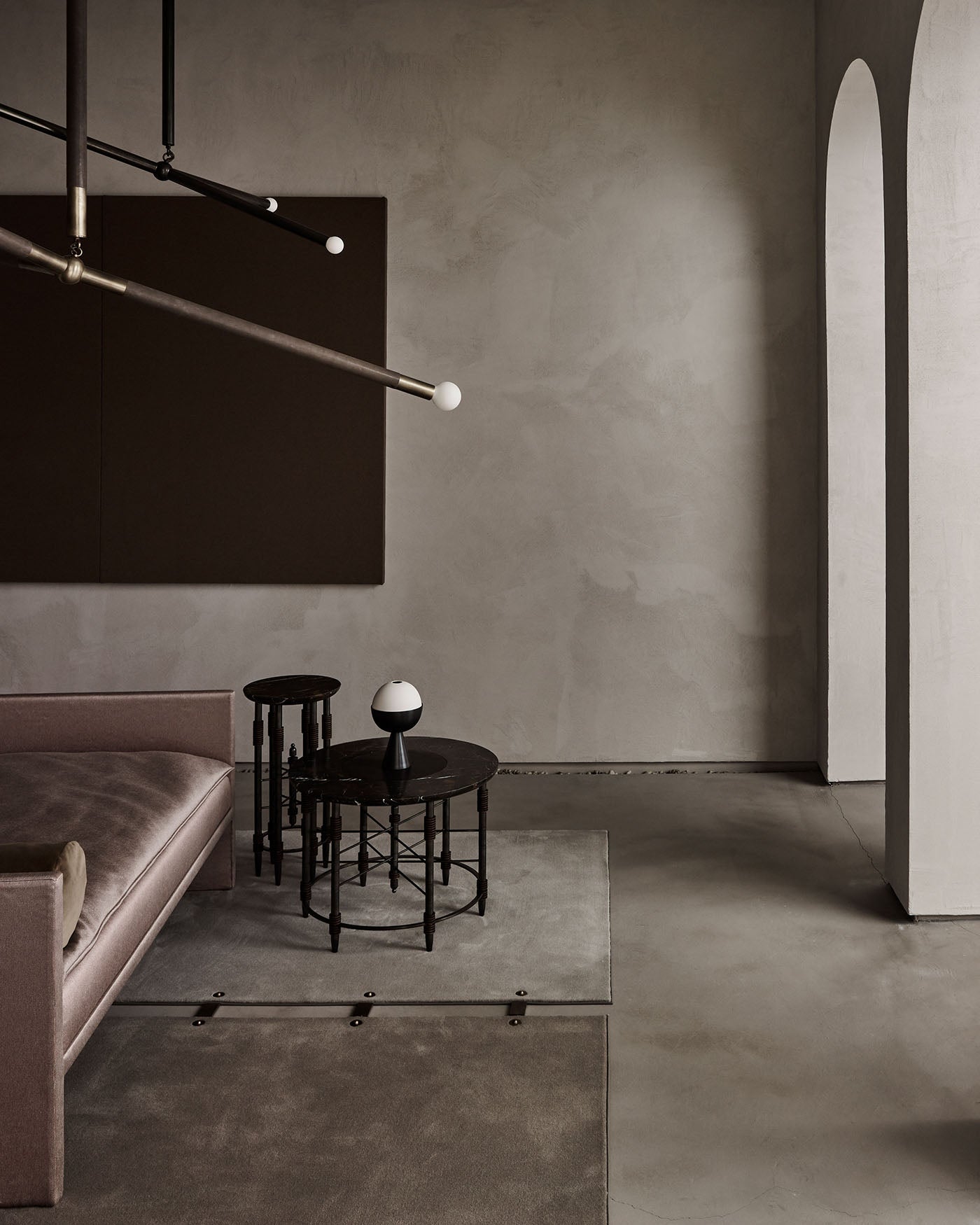Minimalist interior with mauve velvet sofa, nested black metal side tables, modern light fixture, and arched doorway on textured wall