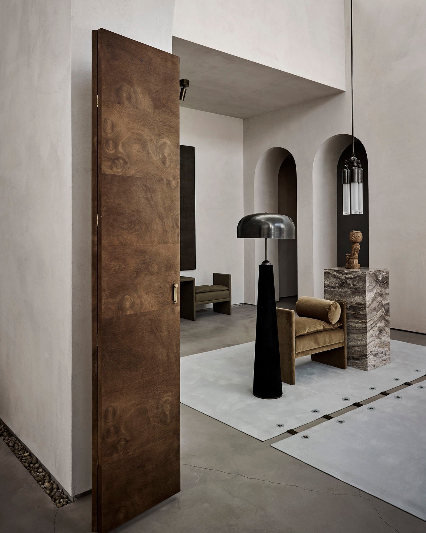 Minimalist interior with a tall wooden door, black floor lamp, velvet armchair, and stone pedestal with sculptural decor