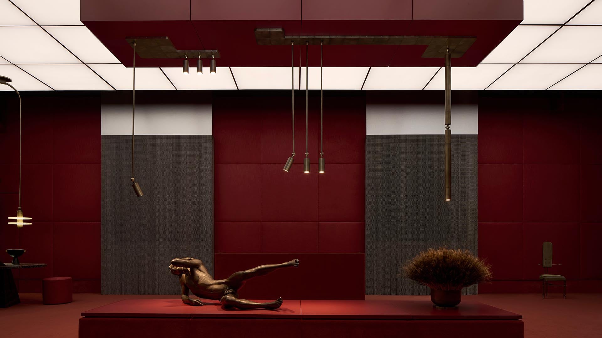 Minimalist interior with deep red walls, suspended brass light fixtures, reclining bronze sculpture, and a dried floral arrangement