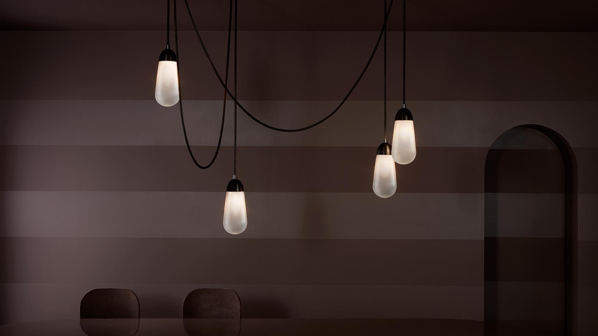 Four elongated pendant lights with warm glow hanging over dark chairs against a striped brown wall with an arched doorway