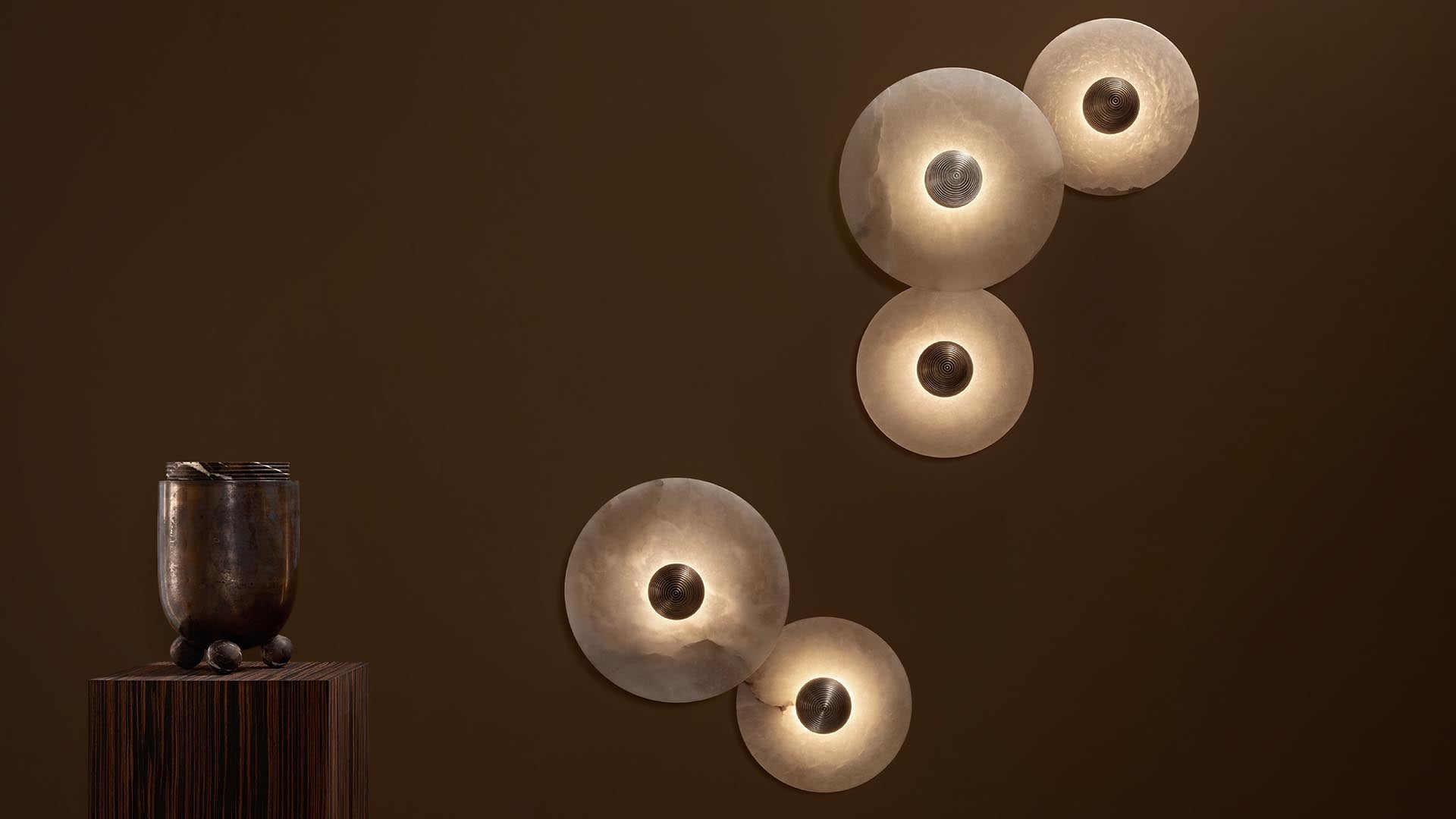 Group of five round pendant lights with textured glass shades glowing against a brown wall above a dark wooden vase on a pedestal