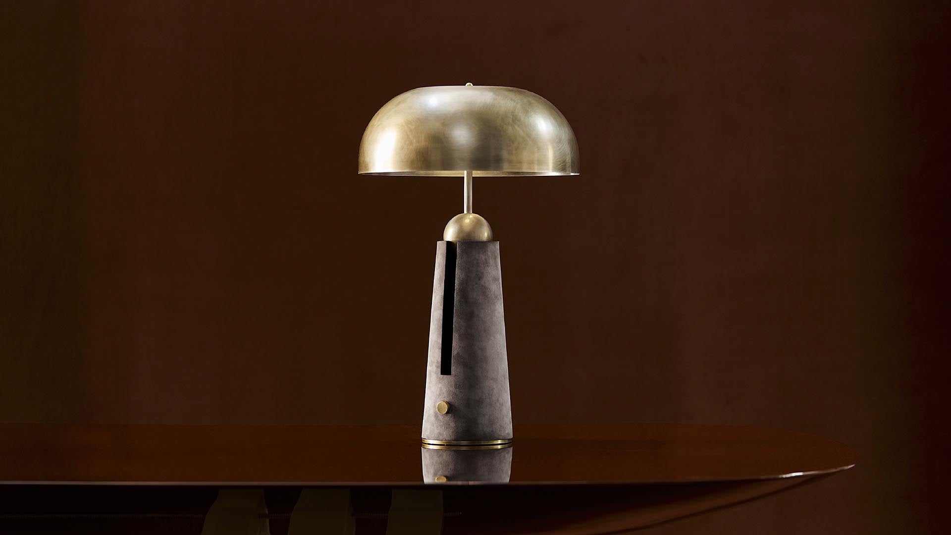 Minimalist table lamp with a brass dome shade and dark stone base on a polished wooden surface against a brown background