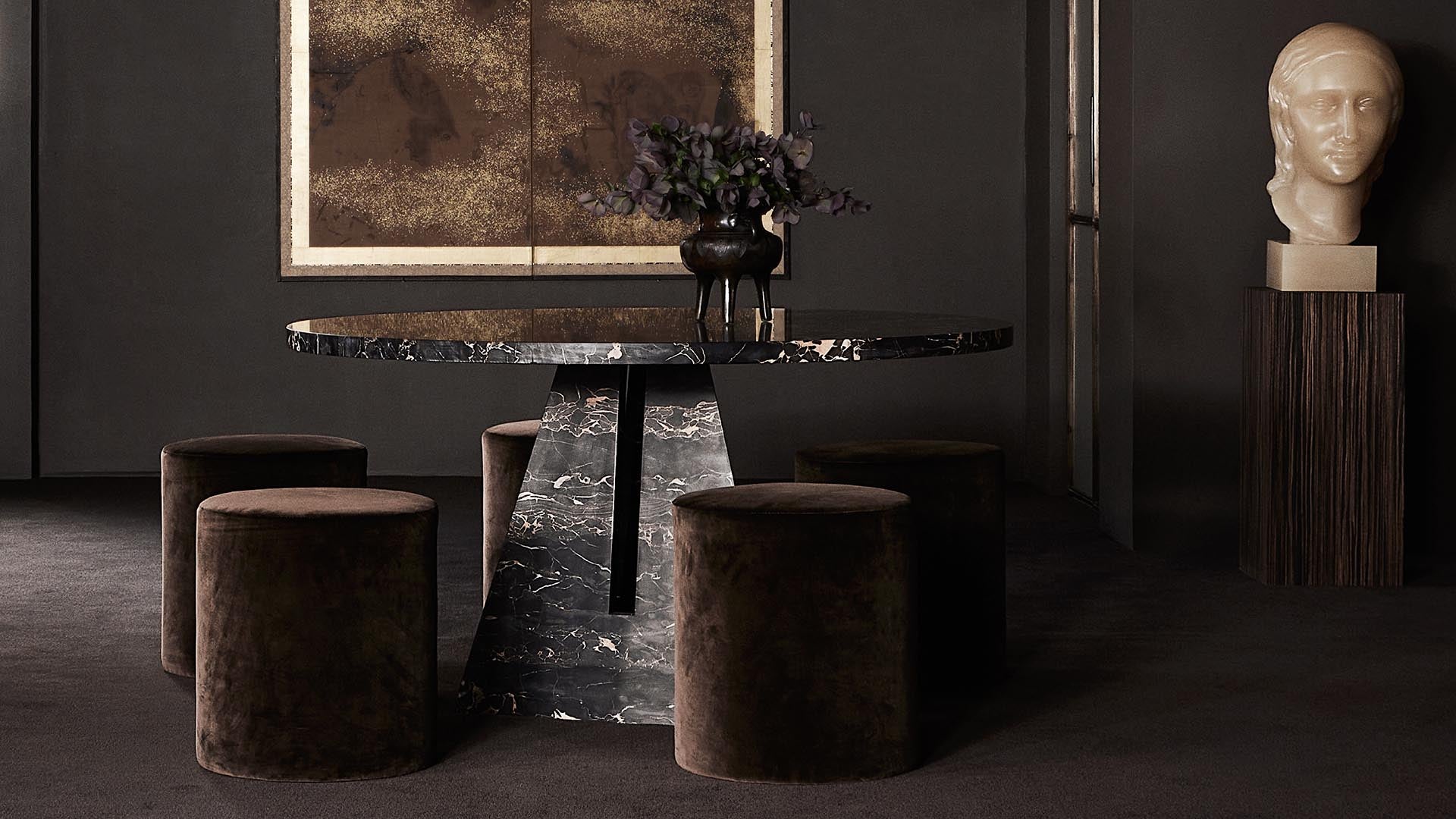 Dark marble round table with black cylindrical base surrounded by brown velvet stools in a dimly lit room with sculpture bust and abstract artwork