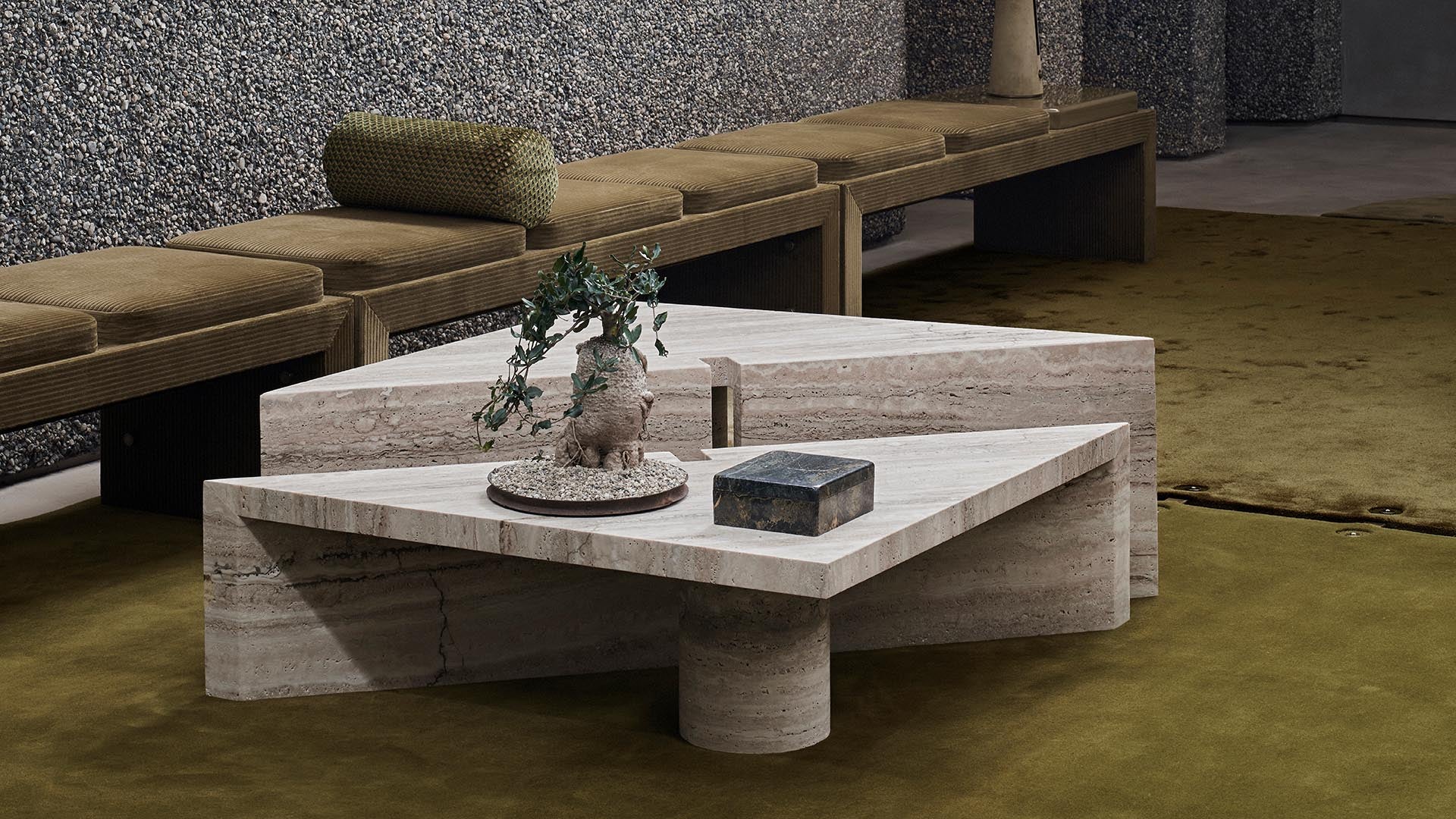 Two triangular stone coffee tables with textured surface on olive green carpet and brown cushioned bench along pebble-textured wall