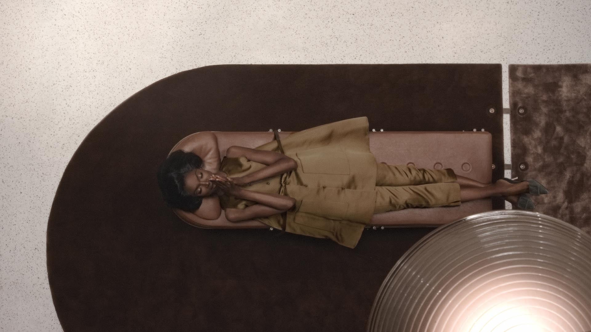 Top-down view of a woman reclining on a pink cushioned bench over a dark curved rug on a light floor.