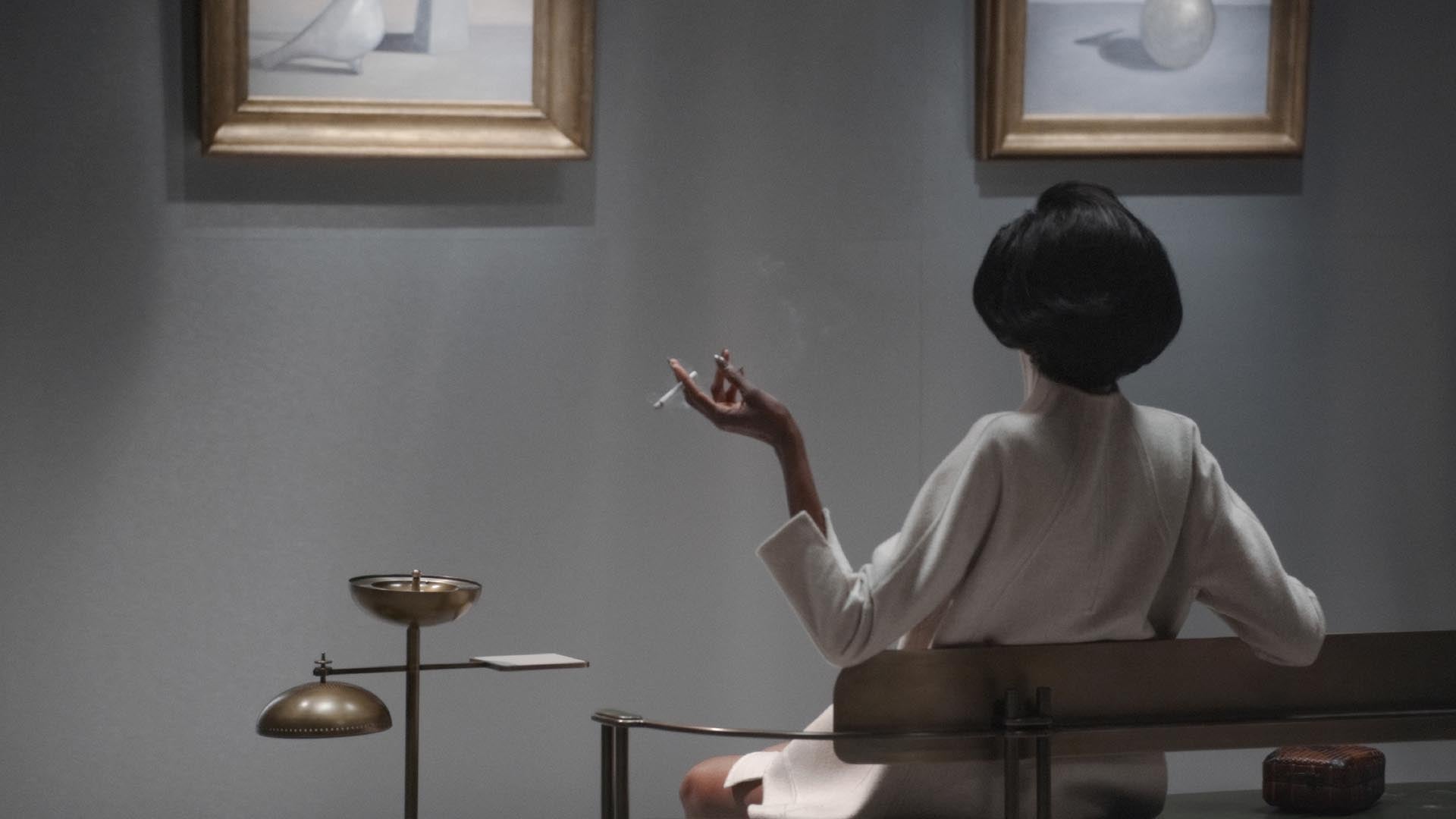 Woman in a white dress sitting on a bench smoking a cigarette under soft wall lighting and framed artwork