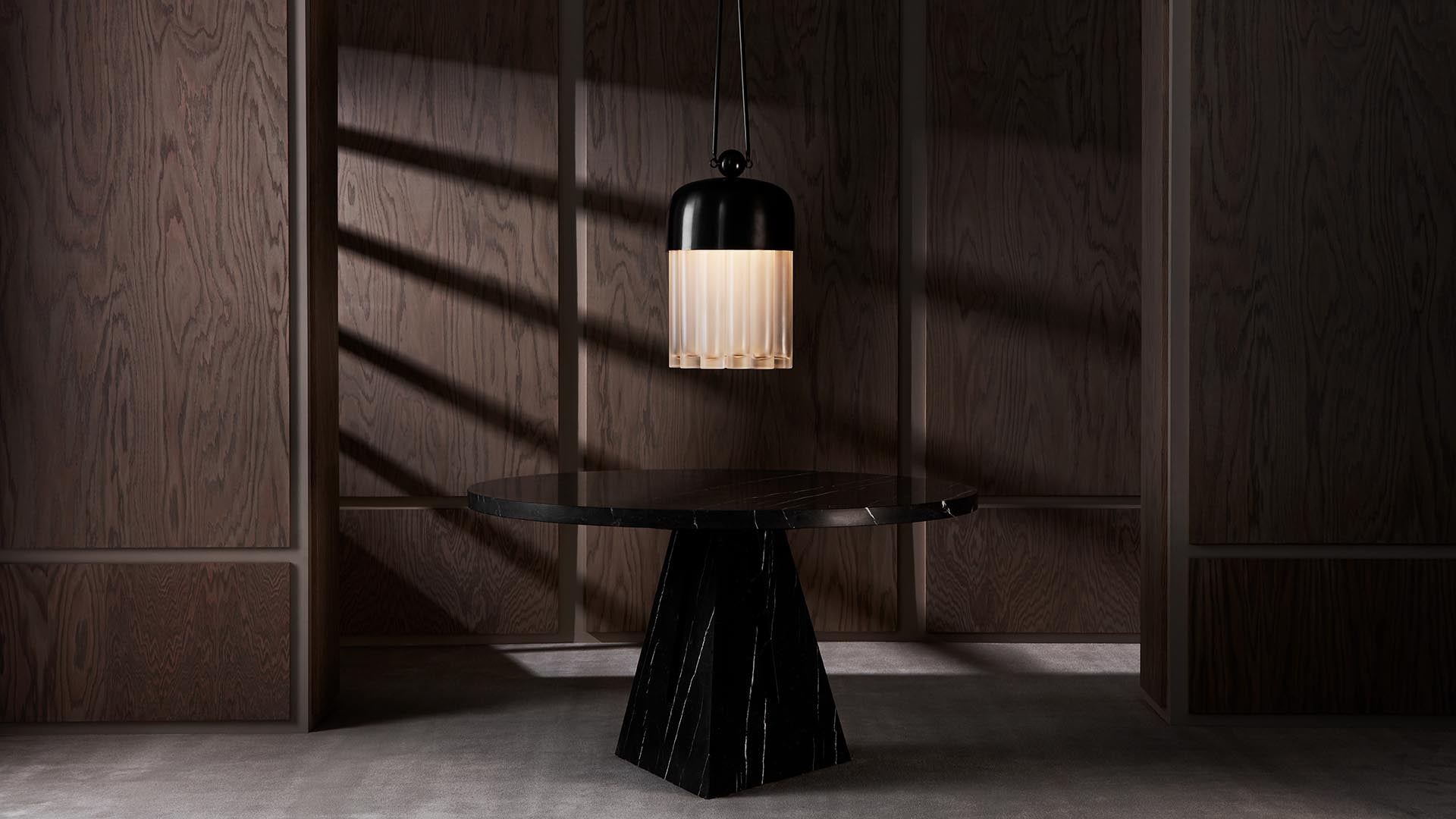 Dark round marble table with a black pendant light above, set against wood-paneled walls casting shadow patterns.