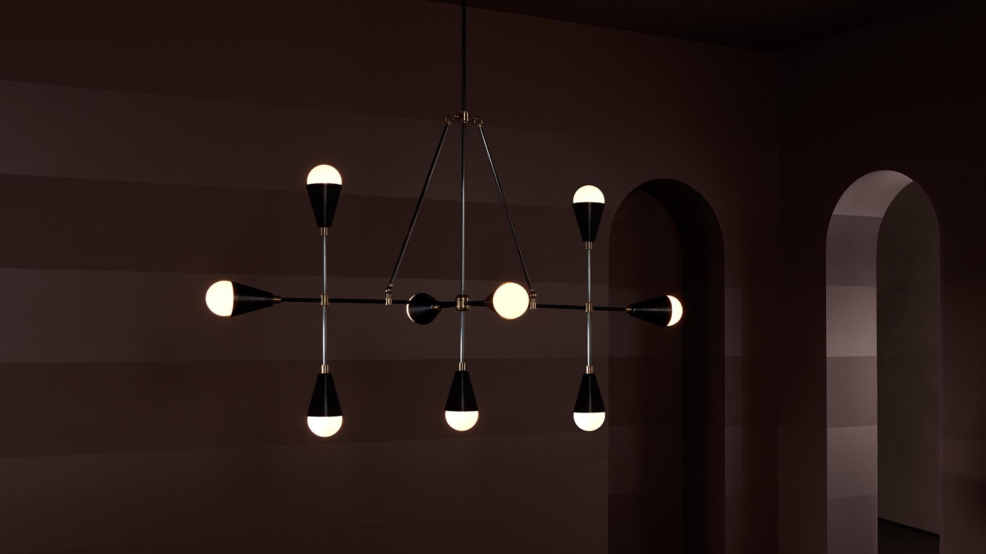 Modern black and brass chandelier with round bulbs suspended in a dimly lit room with striped walls and arched openings
