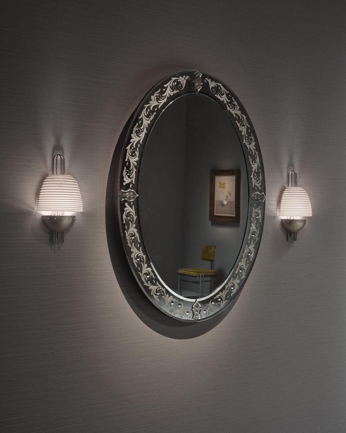 Oval mirror with ornate etched frame flanked by two ribbed glass sconces on textured gray wall