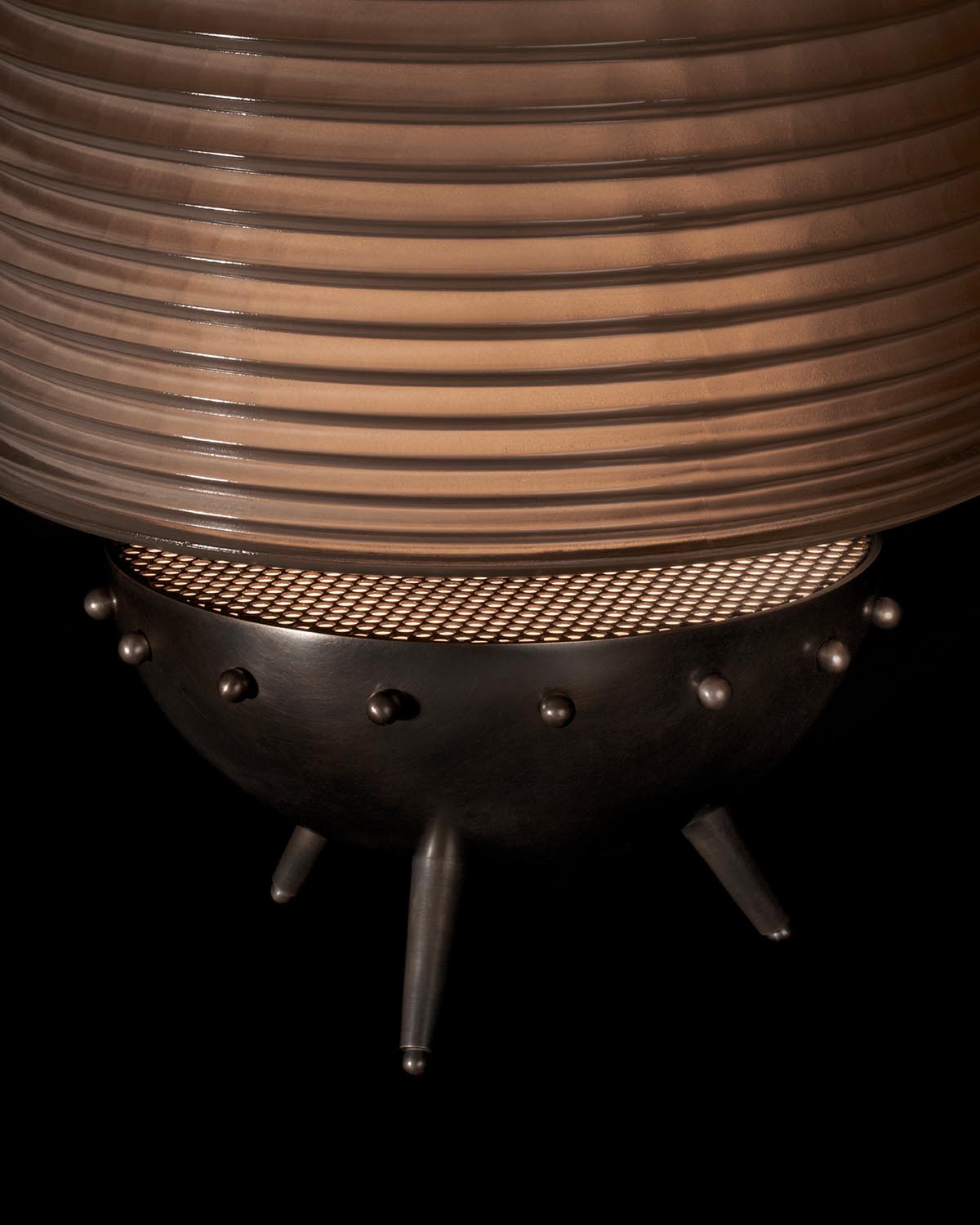 Close-up of SIGNAL X table lamp with ribbed bronze shade and round perforated metal base on three legs against black background