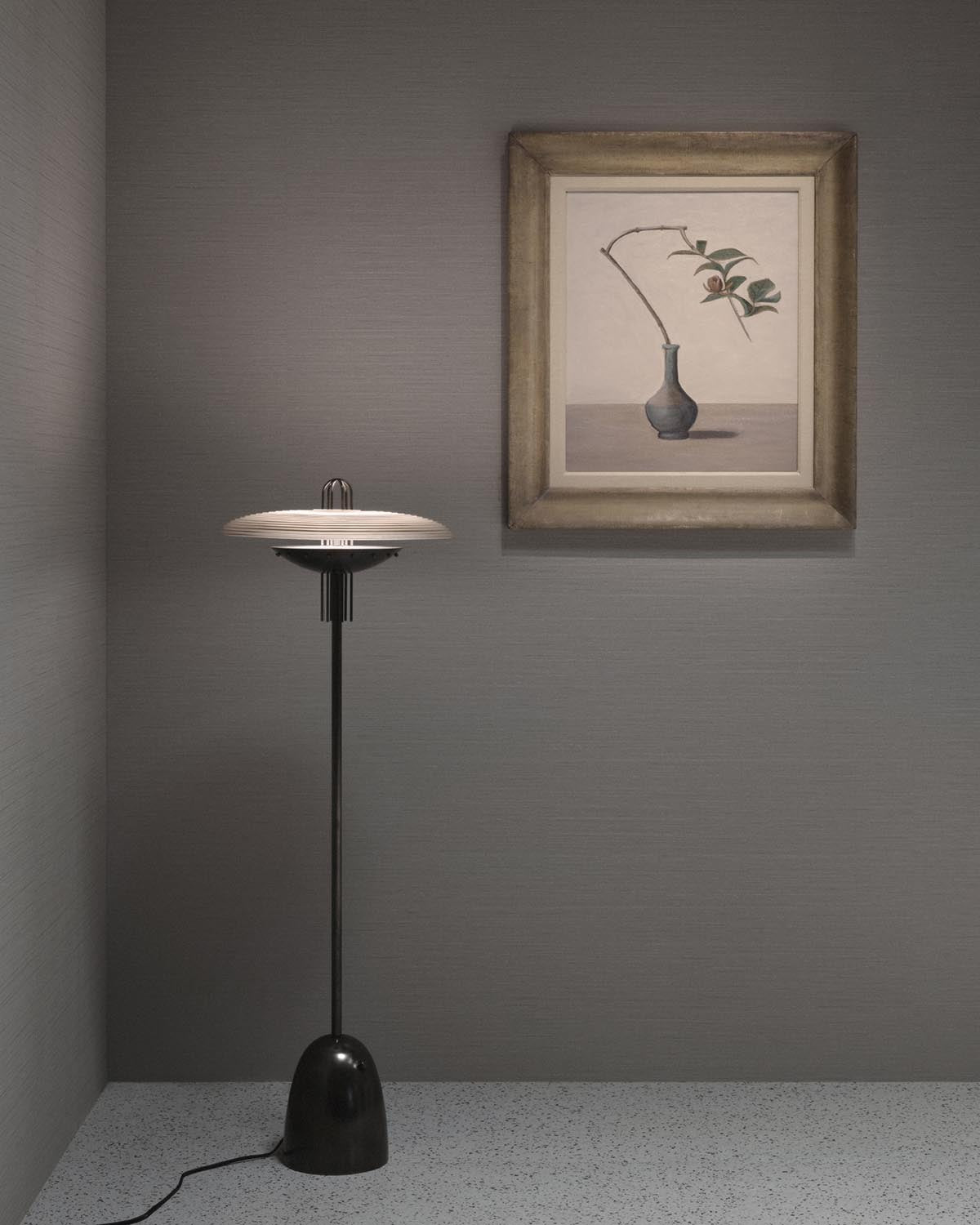 Minimalist floor lamp with black base and white flat circular shade against textured gray walls and framed botanical artwork