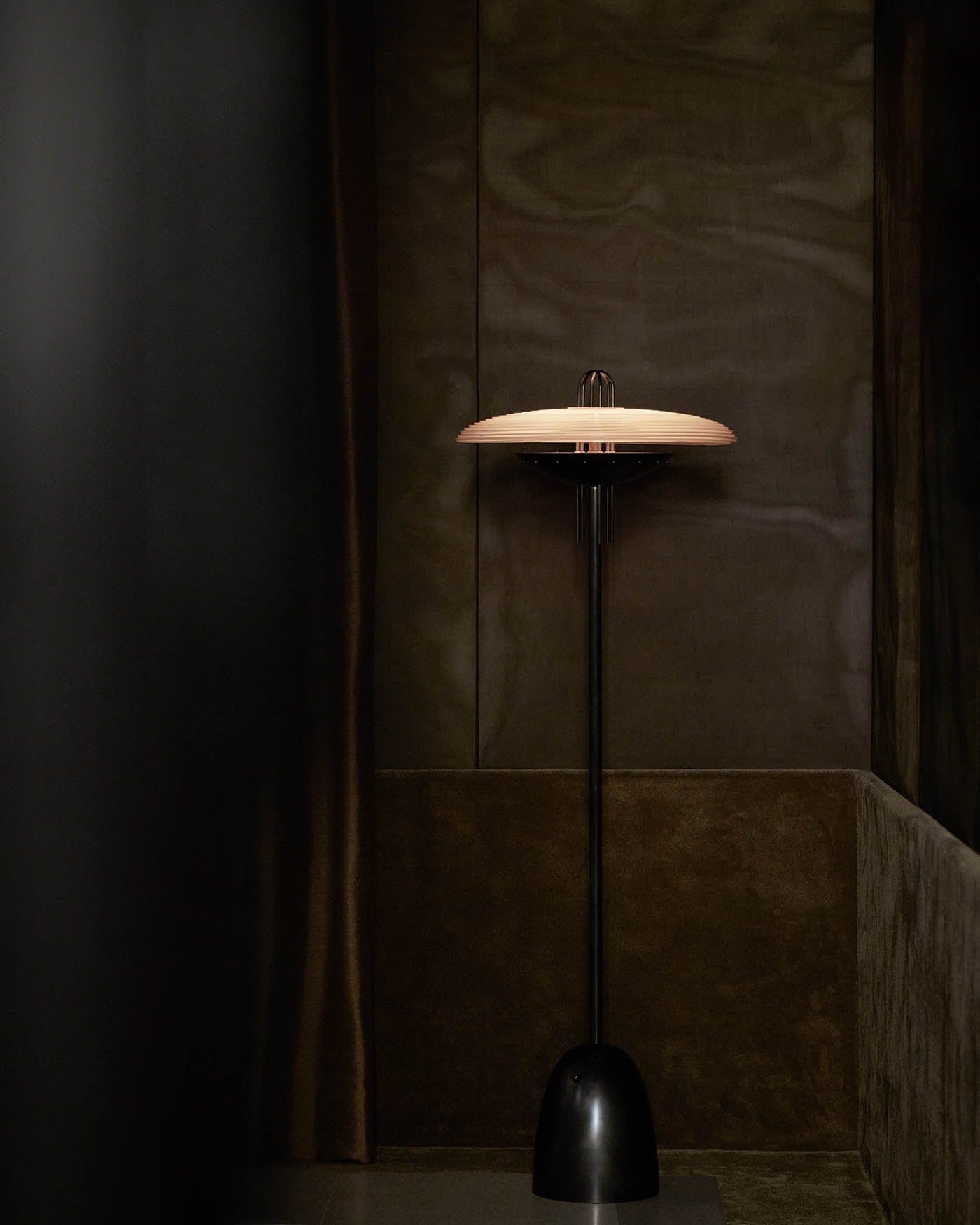 Tall black floor lamp with wide, flat beige lampshade against dark brown textured wall background