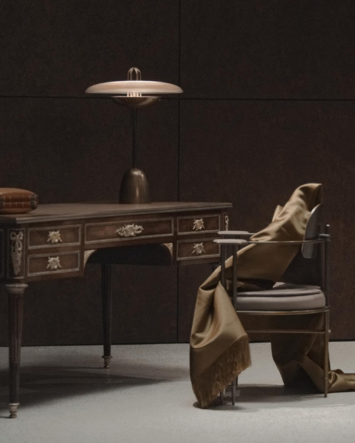 Metallic table lamp with wide circular shade on antique wooden desk surrounded by modern chair draped with brown fabric