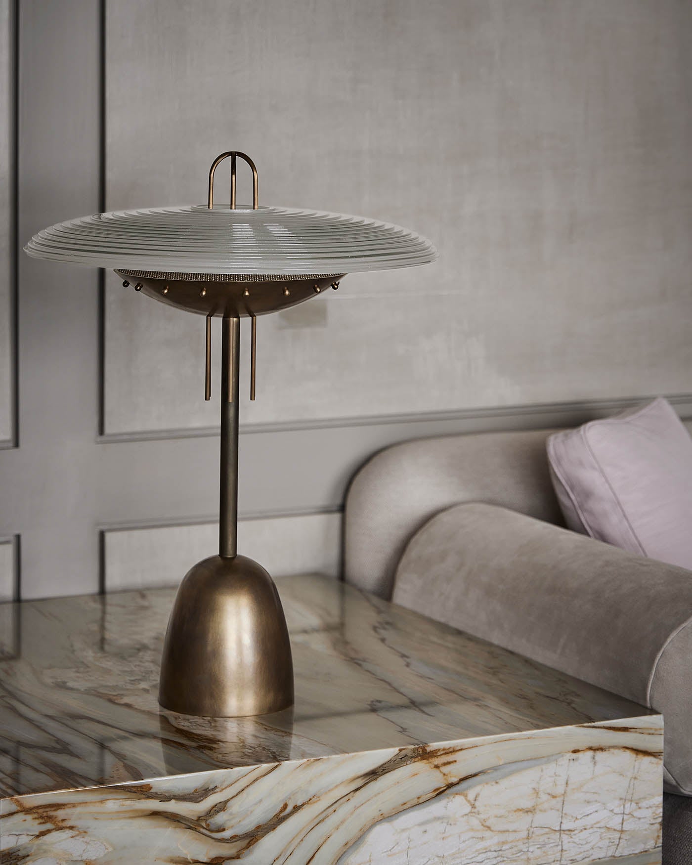 Brass table lamp with a wide ribbed white shade on a polished marble table next to a beige sofa with a pillow