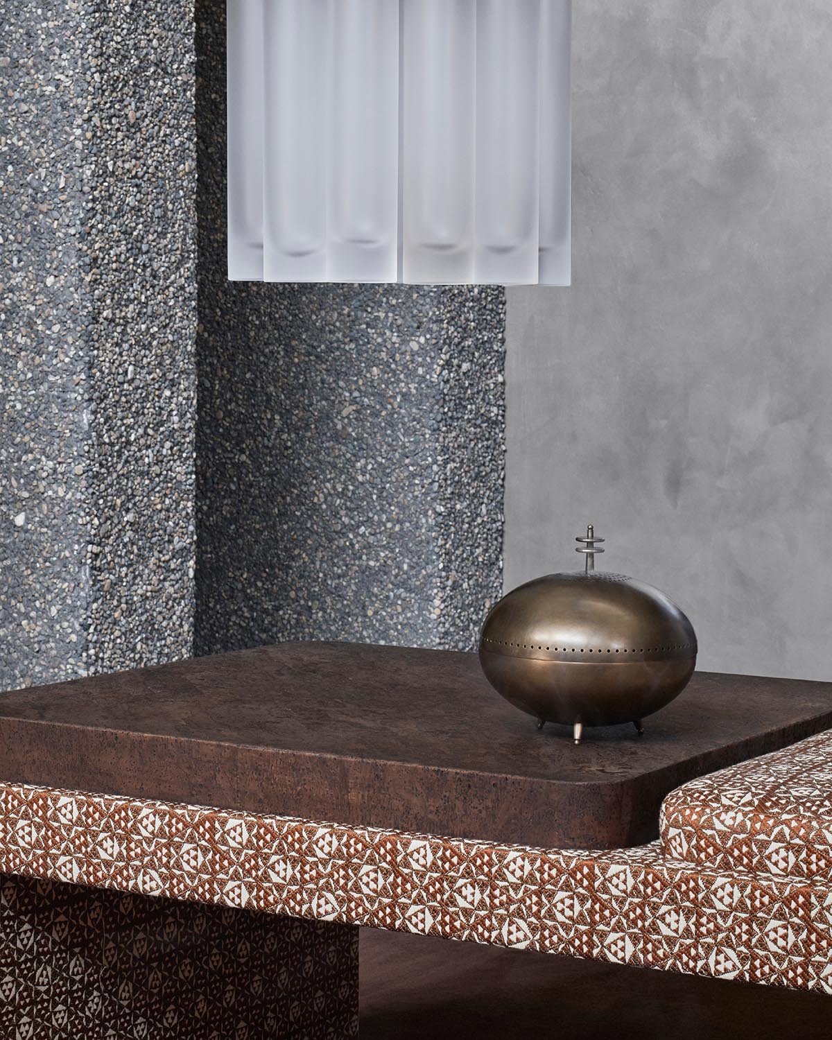 A brass smoking vessel sitting on a wood burl and upholstered table top, beneath a hanging pendant with multiple frosted glass cylinders. 