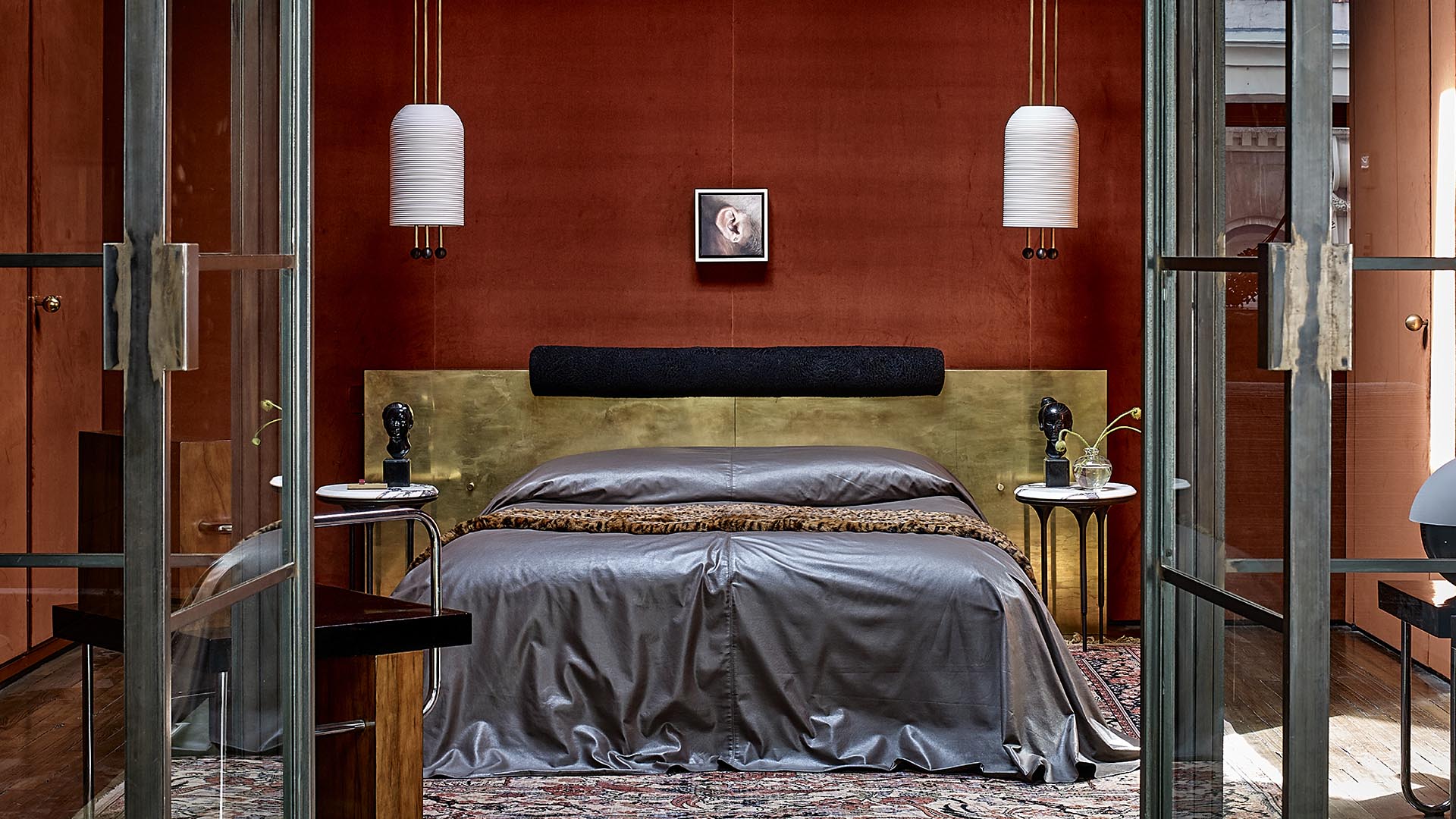 Modern bedroom with a dark gray bedspread, brass headboard, two white pendant lights, and side tables with sculptures and plants