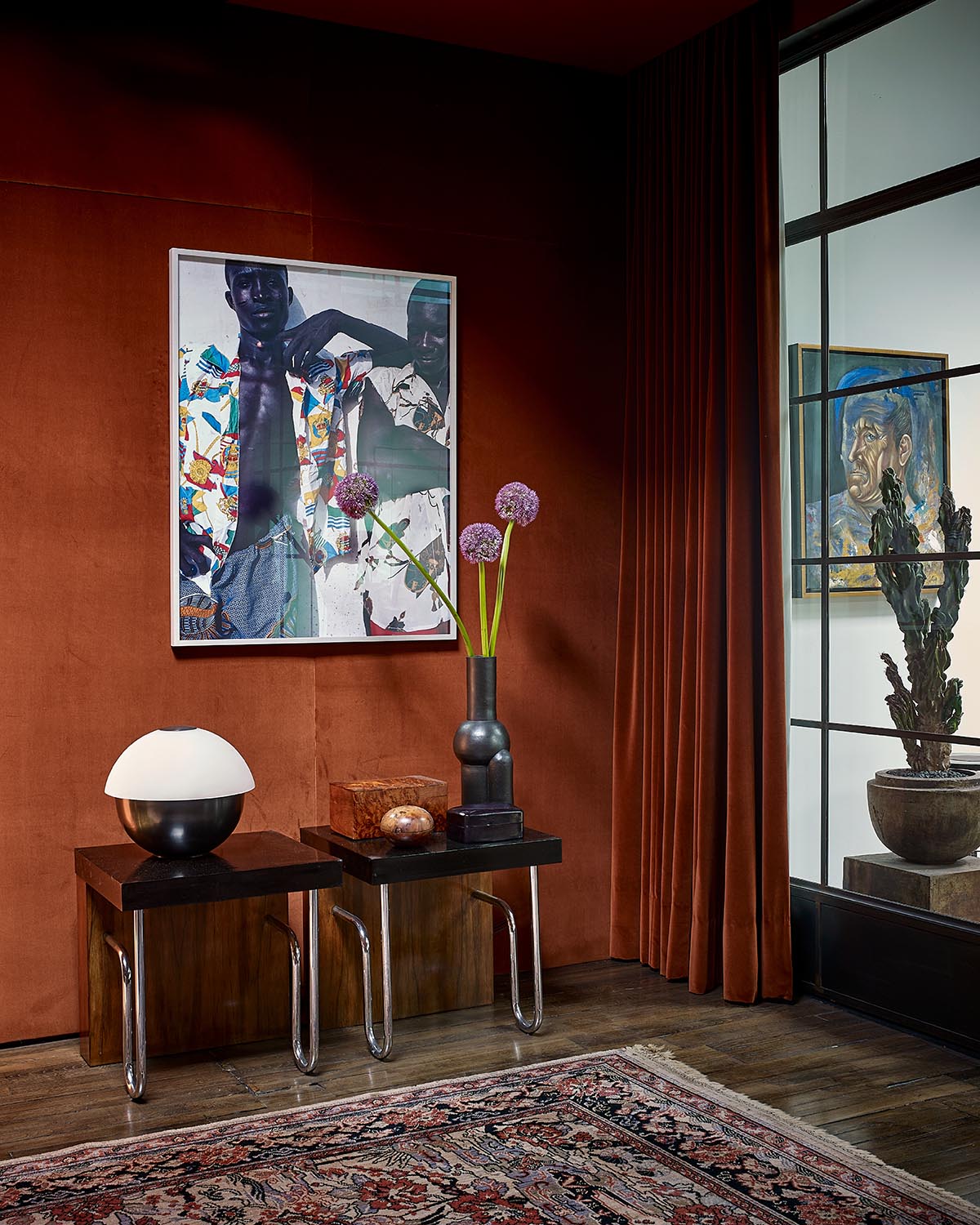 Corner of a room with rust-colored velvet walls and curtains, two black side tables with decor, and framed art on the walls