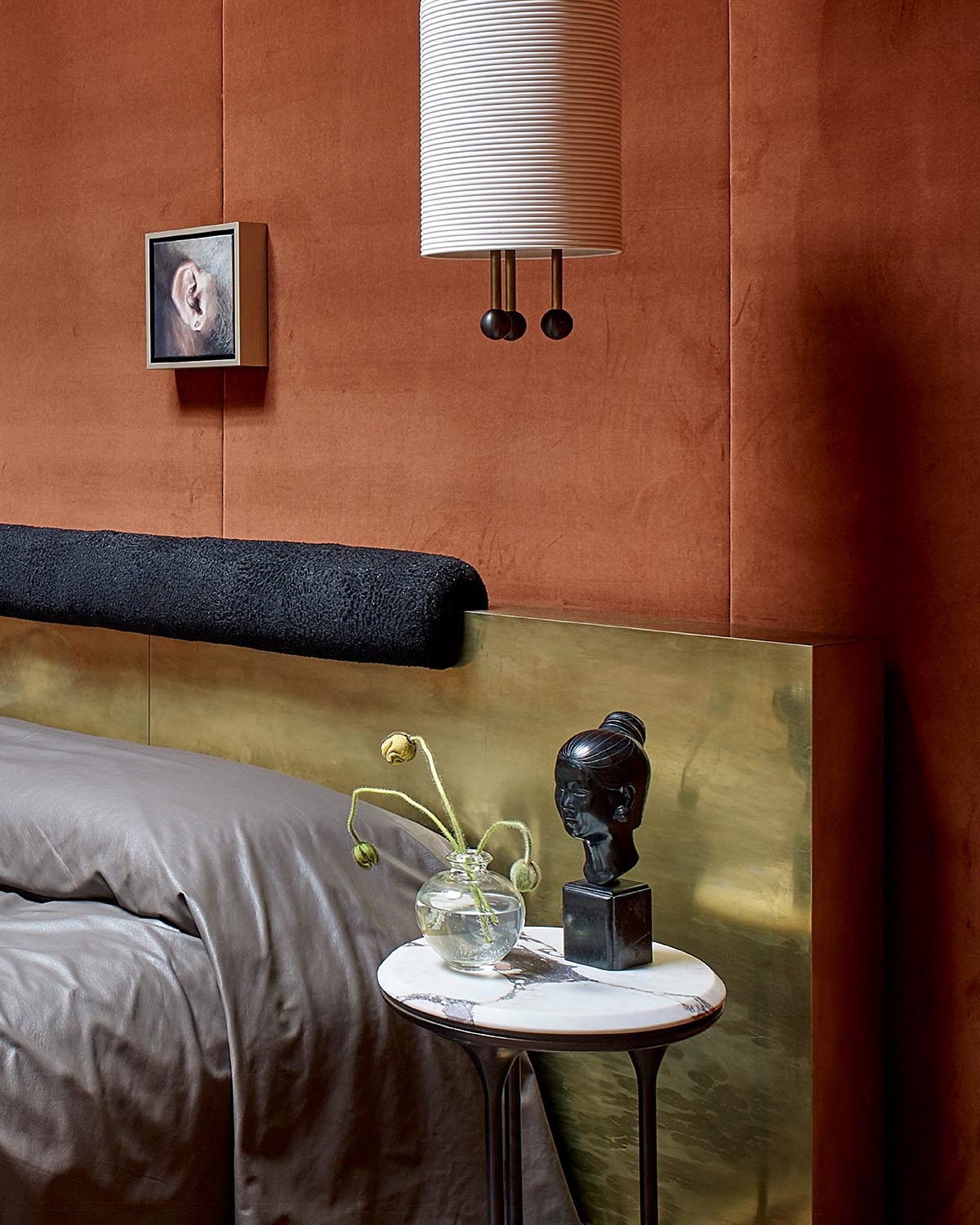 Corner of a bedroom with brown textured walls, brass headboard, grey bedding, marble side table, and decorative sculpture and vase