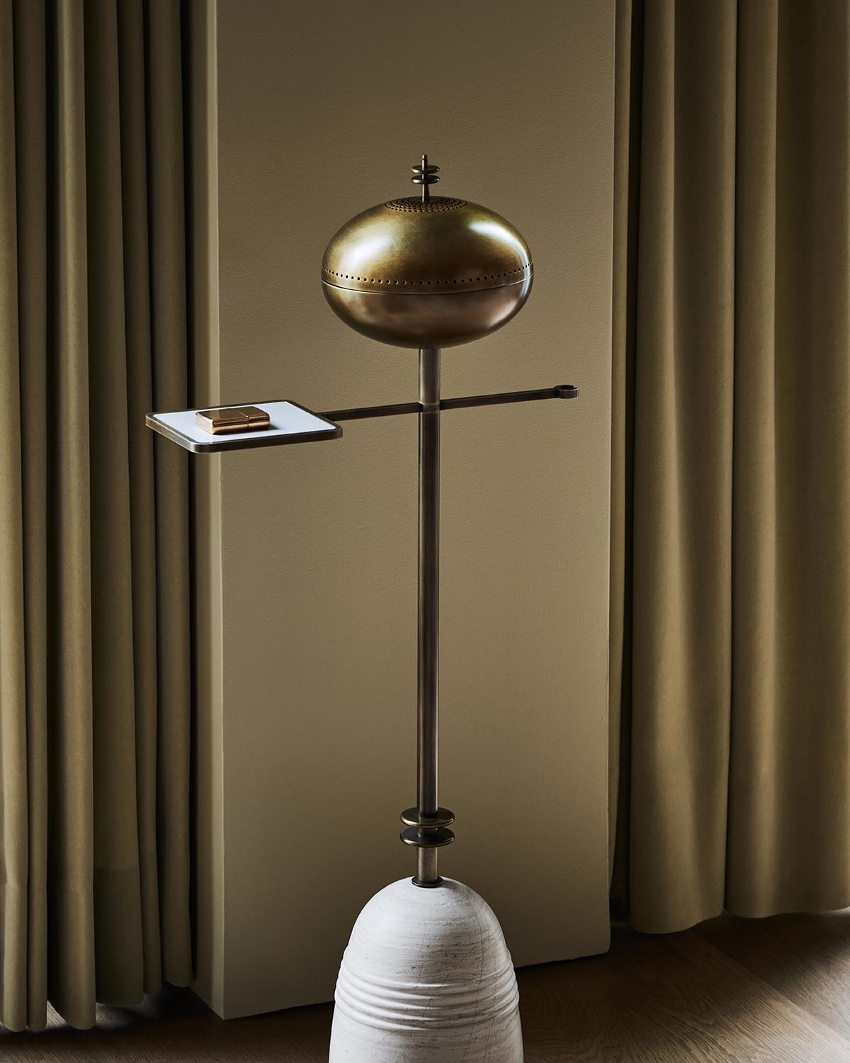 Sculptural floor lamp with a brass oval shade, thin metal arm holding a small white rectangular platform, set against beige curtains.
