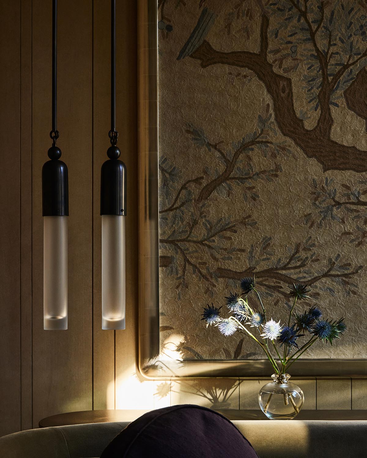Two slim frosted glass pendant lights hanging beside a textured wall mural of a tree branch with a vase of thistle flowers below