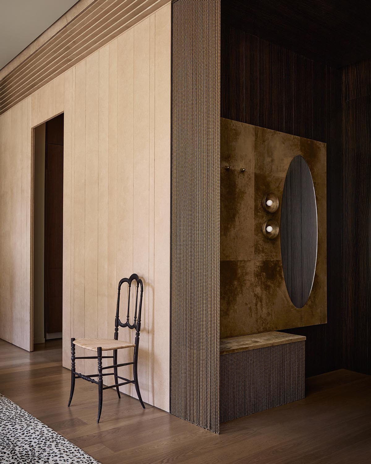 Minimalist interior corner with textured beige walls, dark wooden chair with woven seat, and oval mirror on a brown panel.