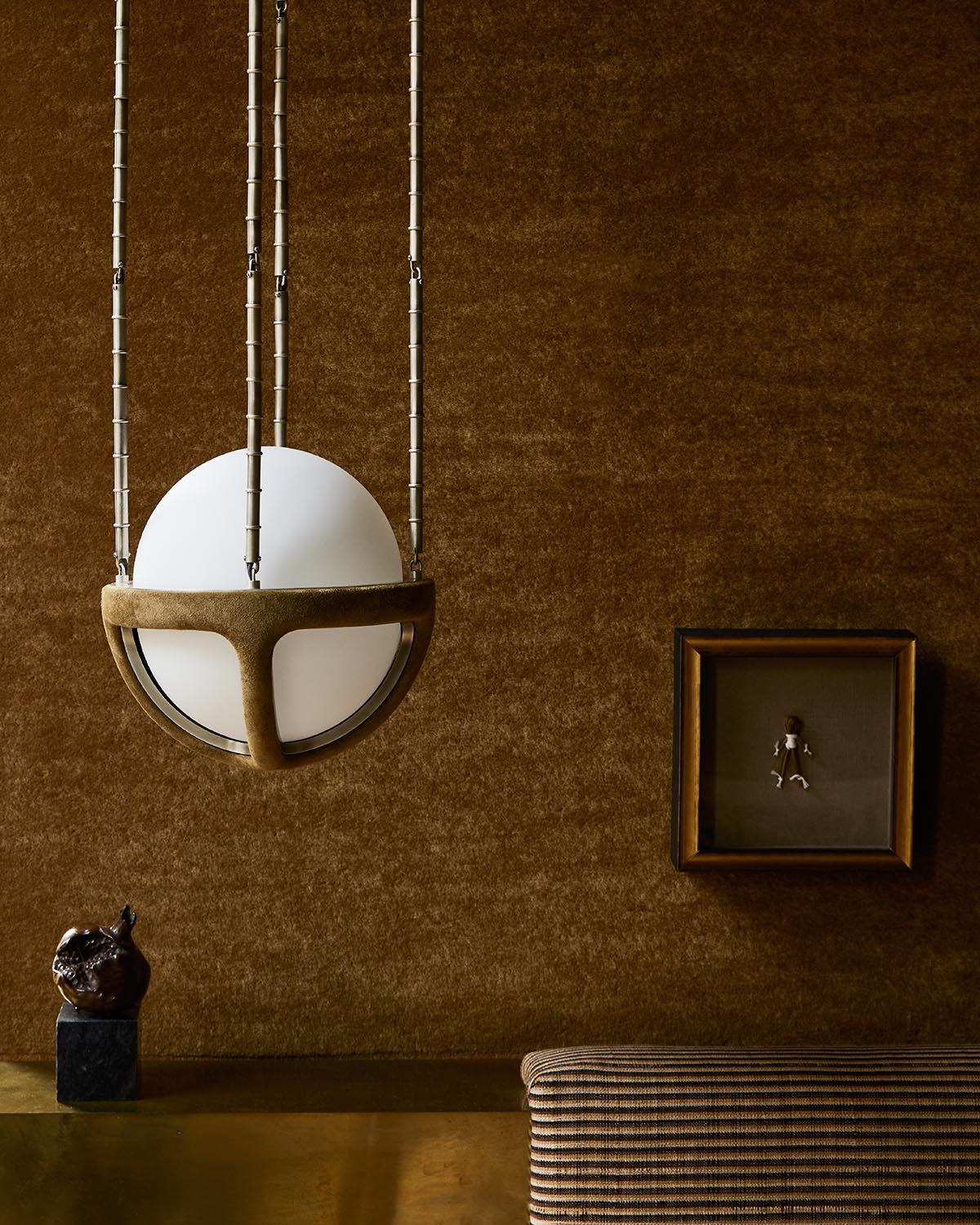 Close-up of a modern spherical pendant light with suede straps against a textured brown wall with framed art and decorative objects