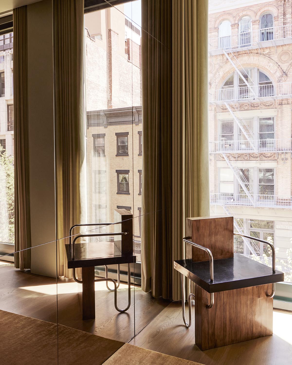 Minimalist wooden chair with metal armrests positioned near large windows with beige curtains and city buildings outside.