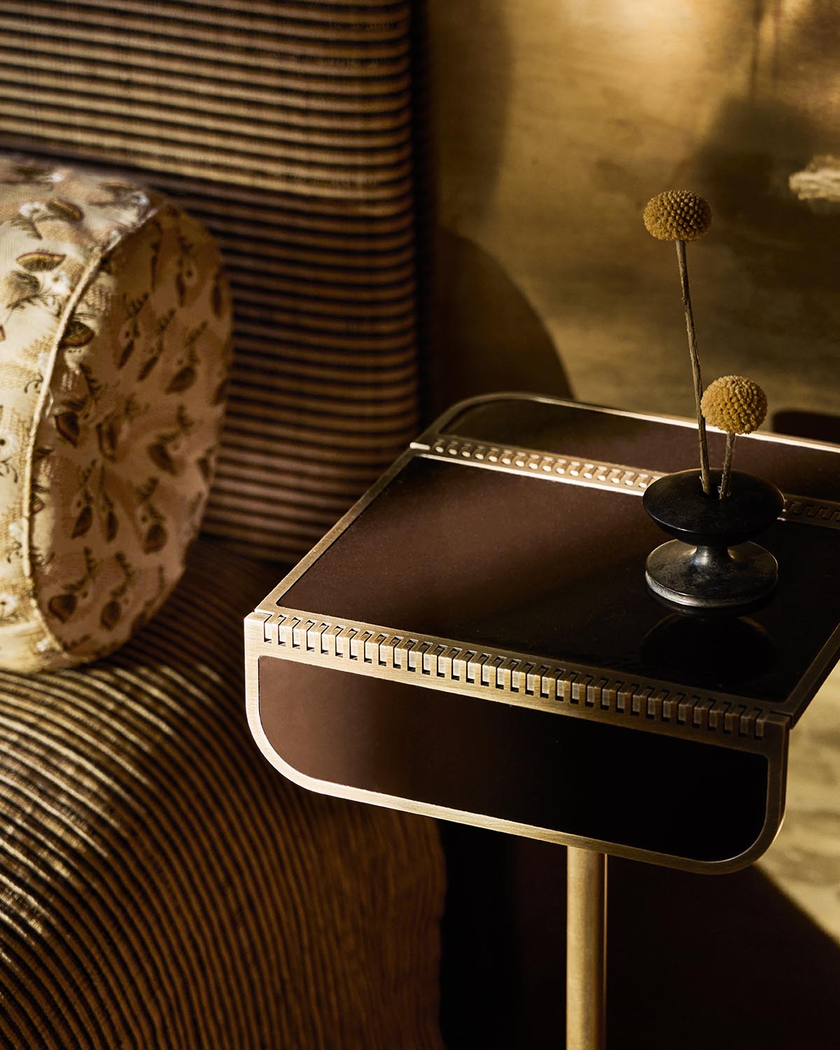 Close-up of a dark brown side table with gold trim and a small black vase holding two dried yellow billy ball flowers