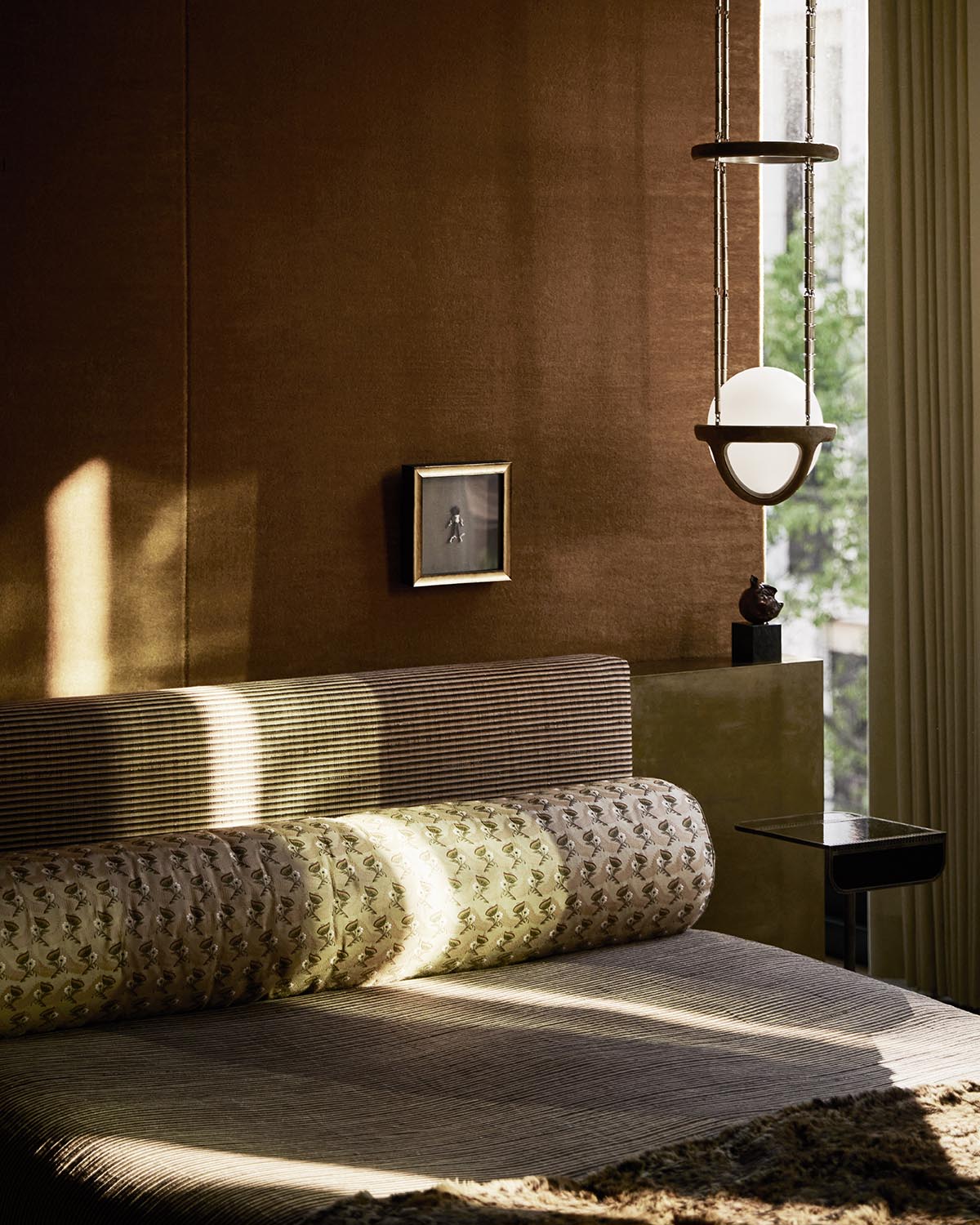 Close-up of a bed with patterned cylindrical pillow and striped headboard against a textured brown wall with soft sunlight.