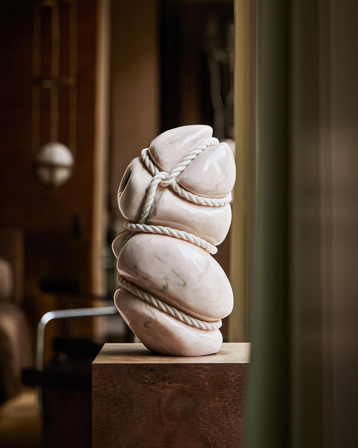 Abstract smooth white ceramic sculpture wrapped in thick rope displayed on a wooden pedestal indoors