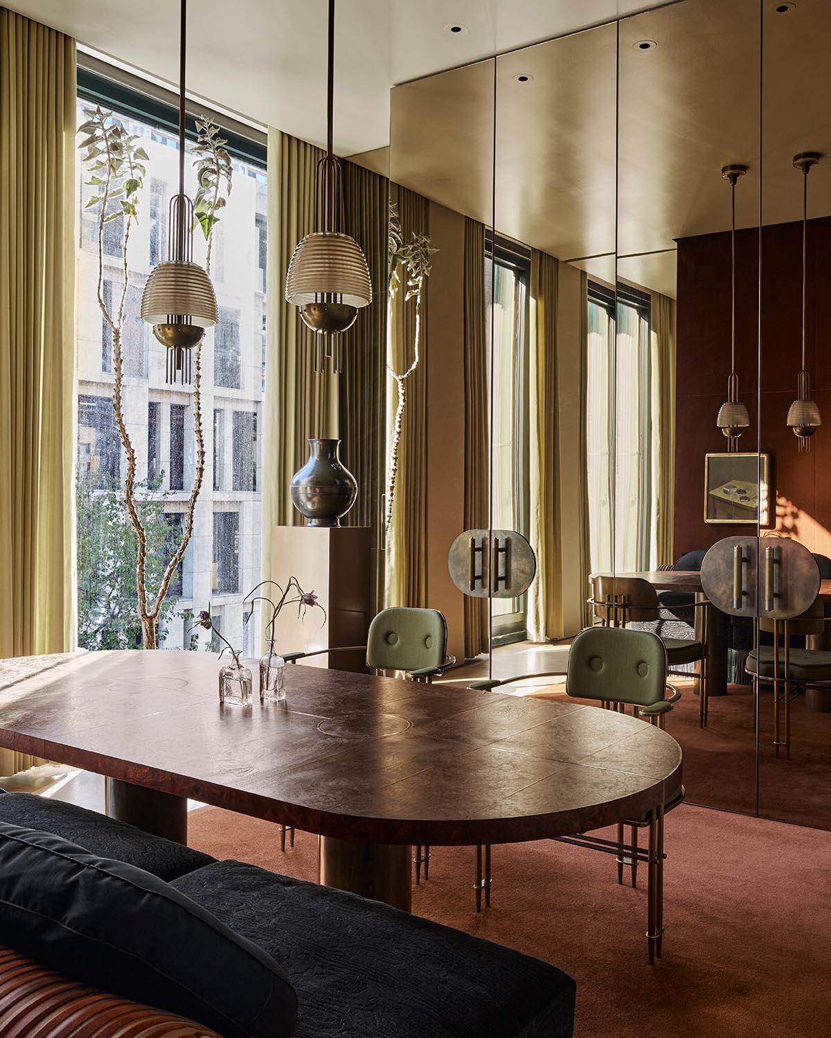 Modern dining room with a large oval wooden table, green chairs, pendant lights, and mirrored wall panels reflecting natural light