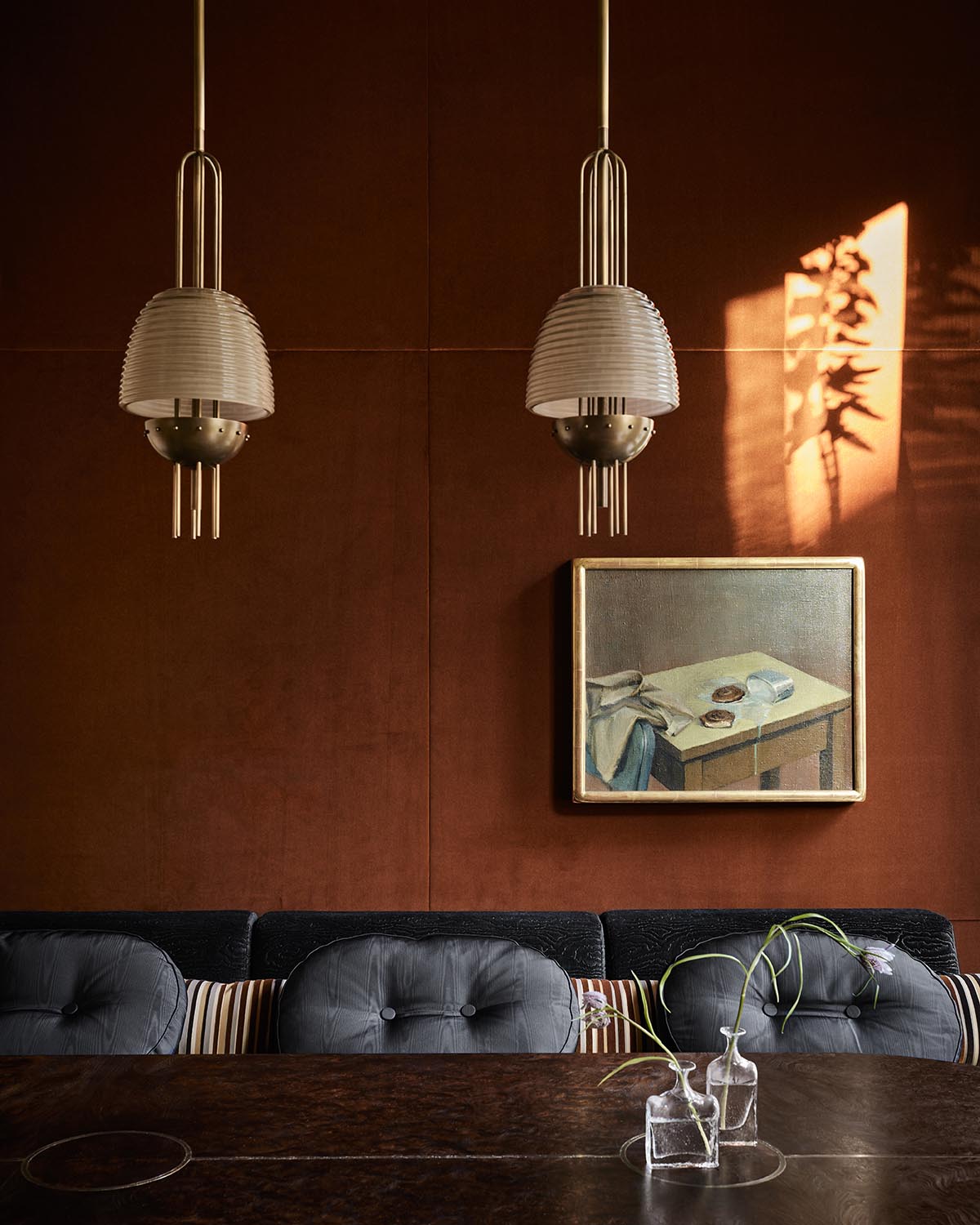 Dark upholstered bench seating with round cushions beneath two pendant lights and a framed painting on a brown wall with sunlight shadow