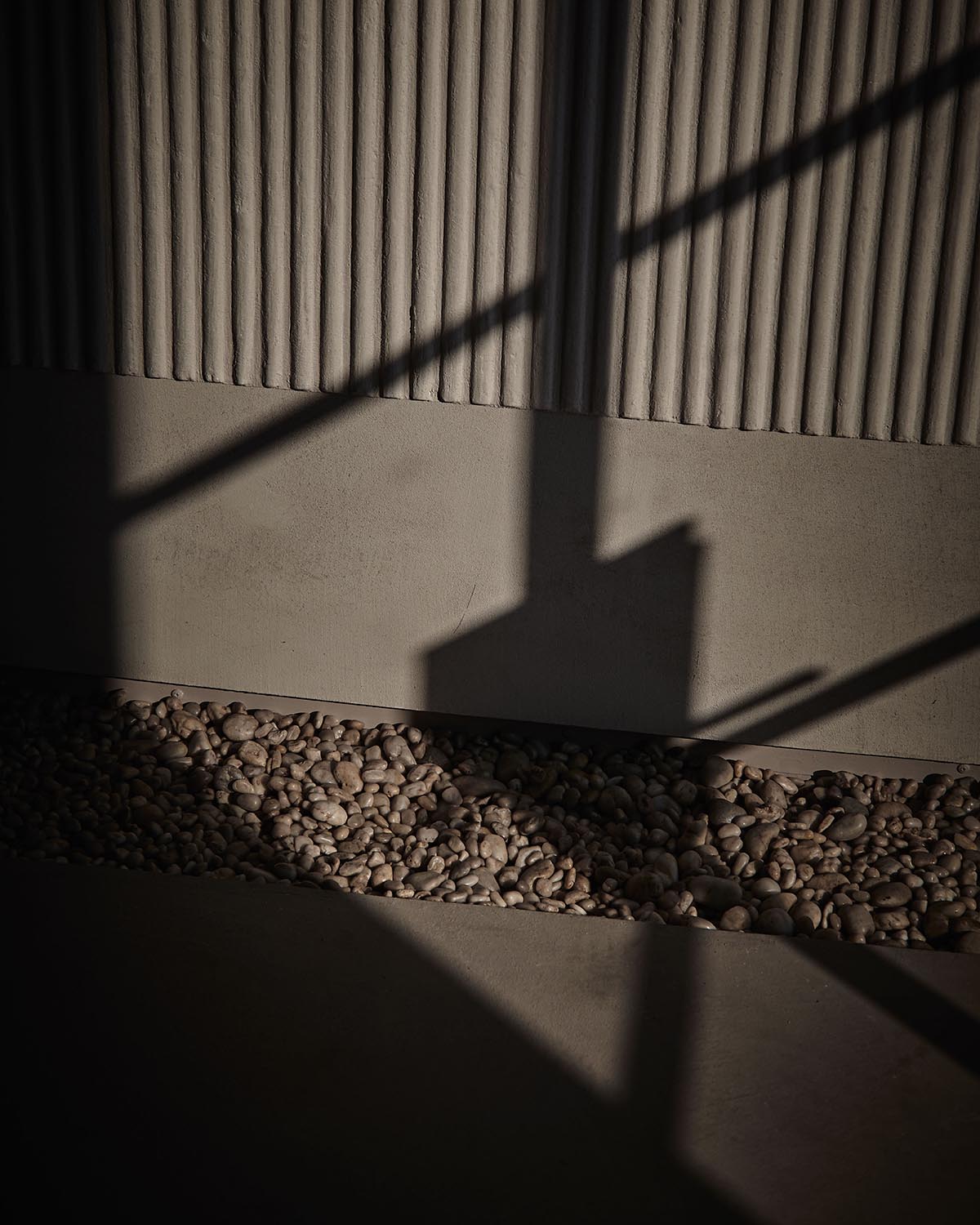 Shadows cast on a textured wall above a narrow bed of smooth river stones on a concrete floor surface