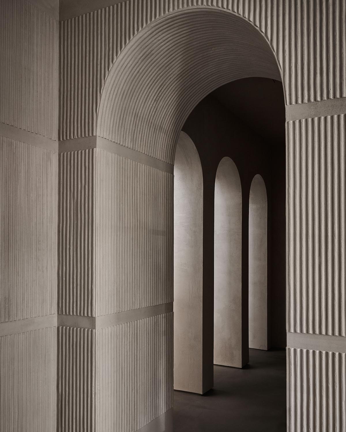 Series of smooth, tall arches with textured vertical ridges on walls creating depth and shadow in a neutral-toned space