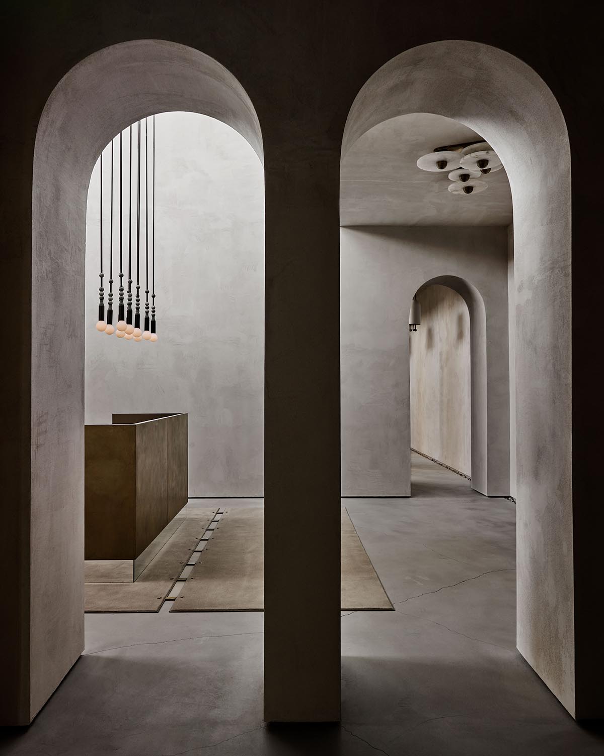Interior view of a minimalist space with arched doorways, hanging pendant lights, and textured neutral-toned walls and floors