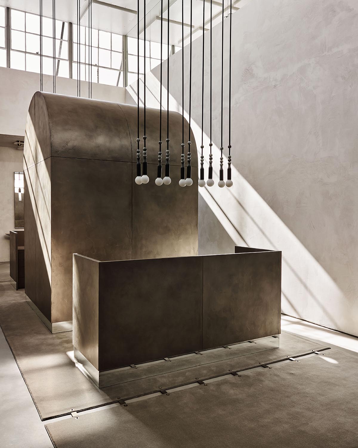 Minimalist interior with curved concrete structure, hanging pendant lights in a row, and sunlight casting shadows on textured walls