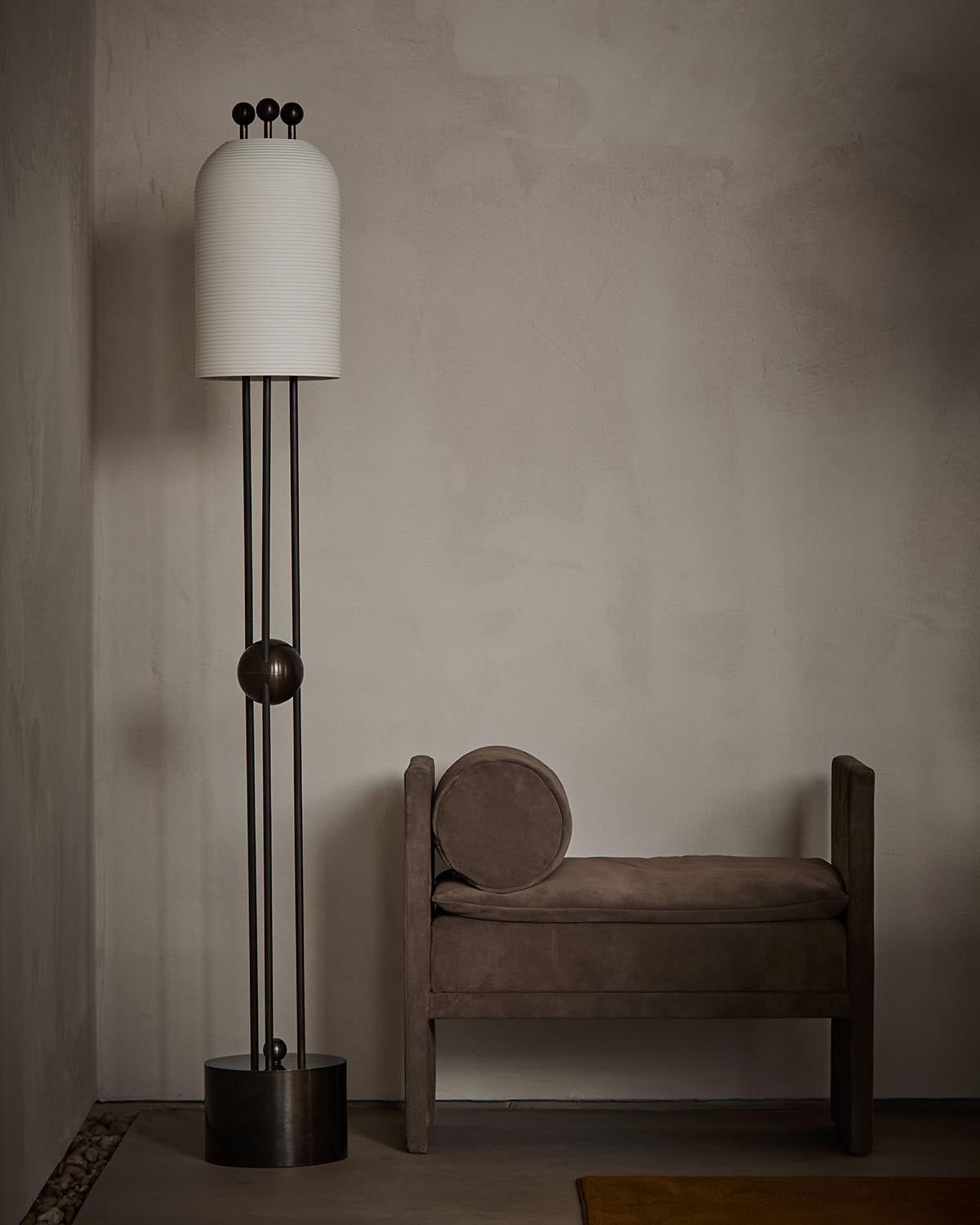 Tall floor lamp with white cylindrical shade and dark metal frame next to a brown suede bench with round cushion against a textured wall