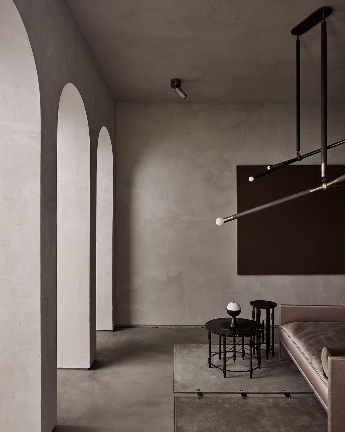 Minimalist interior with arched wall openings, concrete floor, dark round tables, and a muted sofa under modern lighting fixture