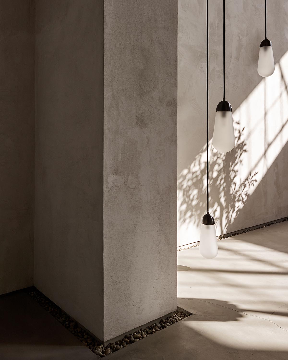 Minimalist interior corner with textured beige walls, pendant lights, and shadows of plants cast by sunlight.