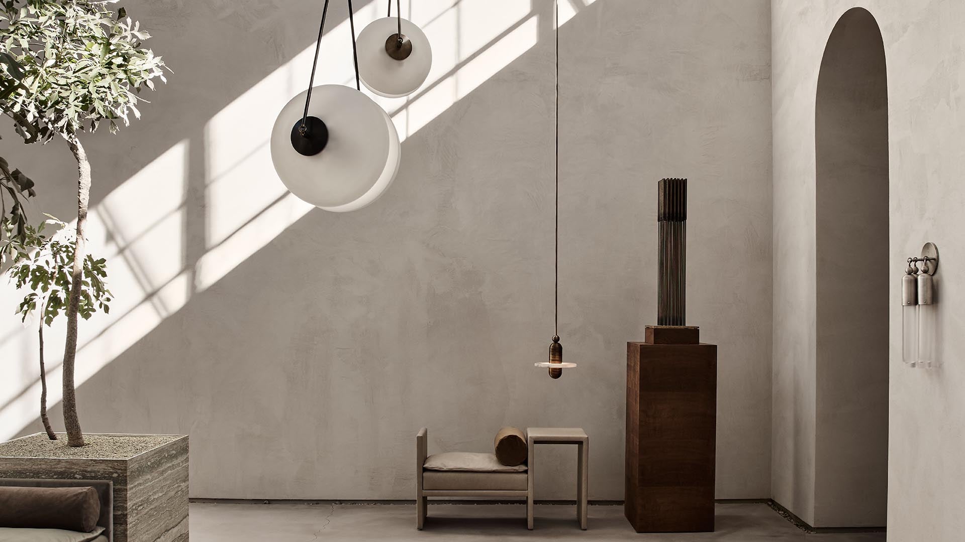 Minimalist interior with textured beige walls, pendant lights, potted tree, and simple wooden chair with ottoman