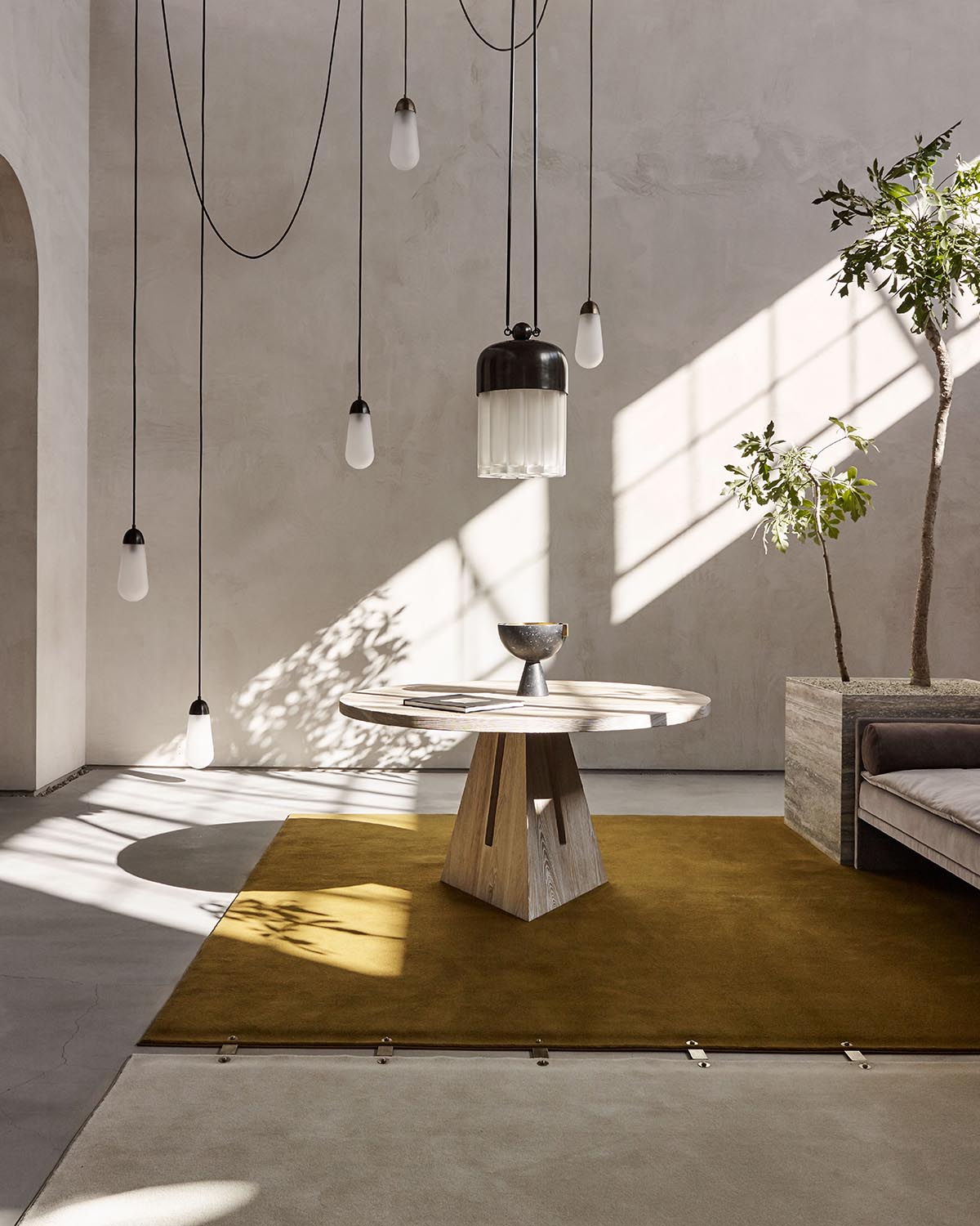 Minimalist interior with round wooden table on mustard rug, pendant lights hanging from ceiling, and potted plant by window shadow