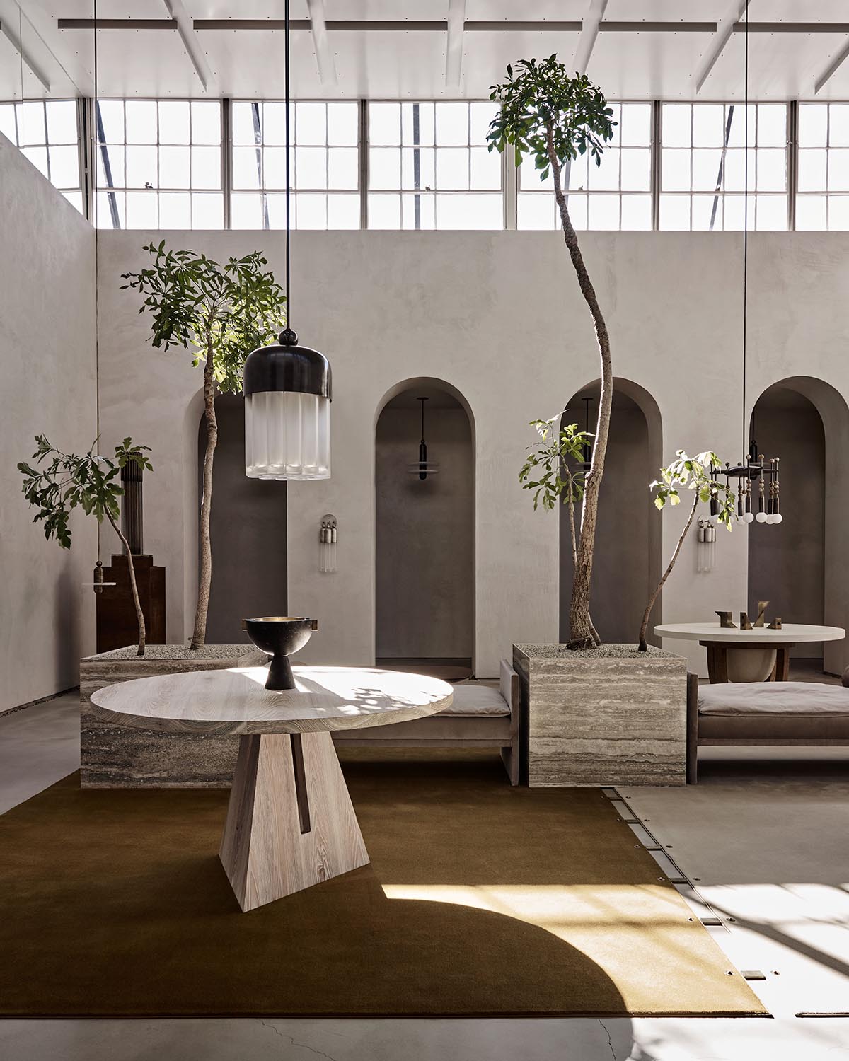 Minimalist interior with round wooden table, built-in stone planters holding tall trees, and large windows overhead