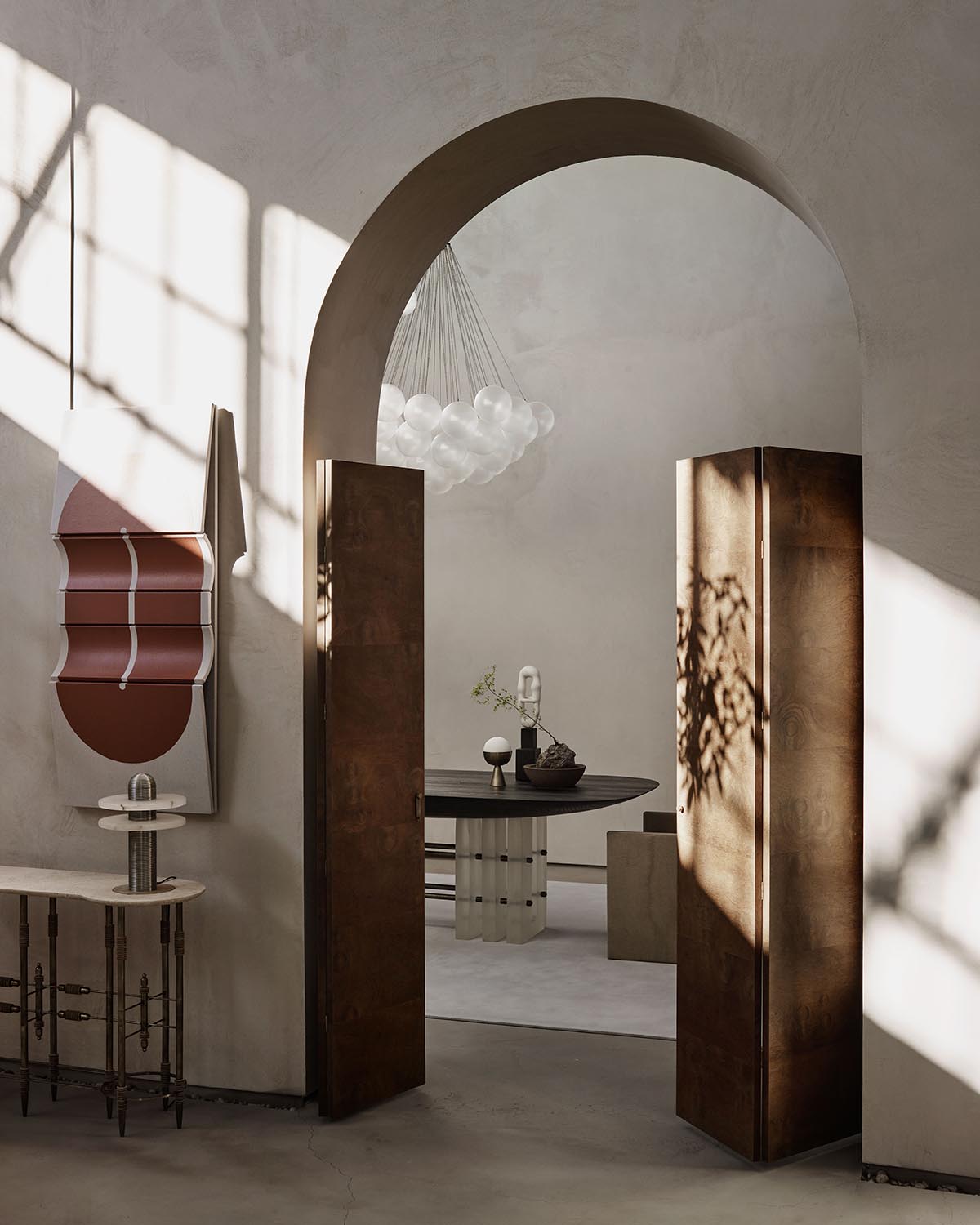 Sunlit arched doorway with tall rust-colored metal doors opening into a room with round black table and sculptural light fixture