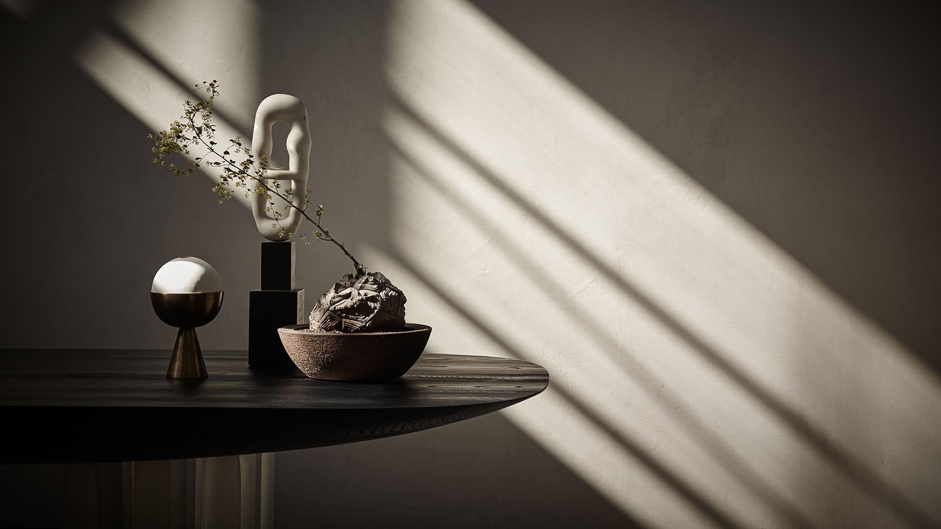 Minimalist dark wooden table with sculptural ceramic objects and delicate dried branch casting shadows on textured wall
