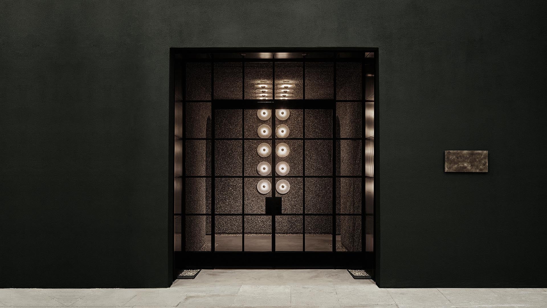 Dimly lit interior space viewed through a large grid-style glass window with circular lights on a dark textured wall inside