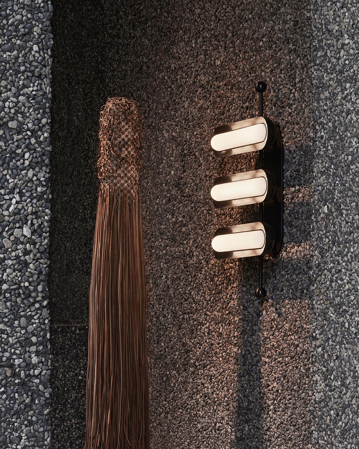 Modern wall light fixture with three elongated bulbs mounted on textured dark gray stone wall beside long hanging woven decor