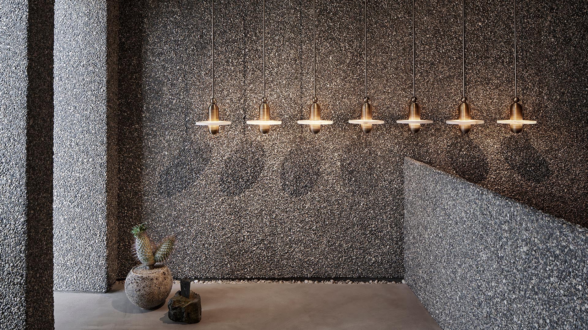 Row of seven brass pendant lights casting shadows on a textured gray pebble wall above a concrete floor with two rustic pottery pieces