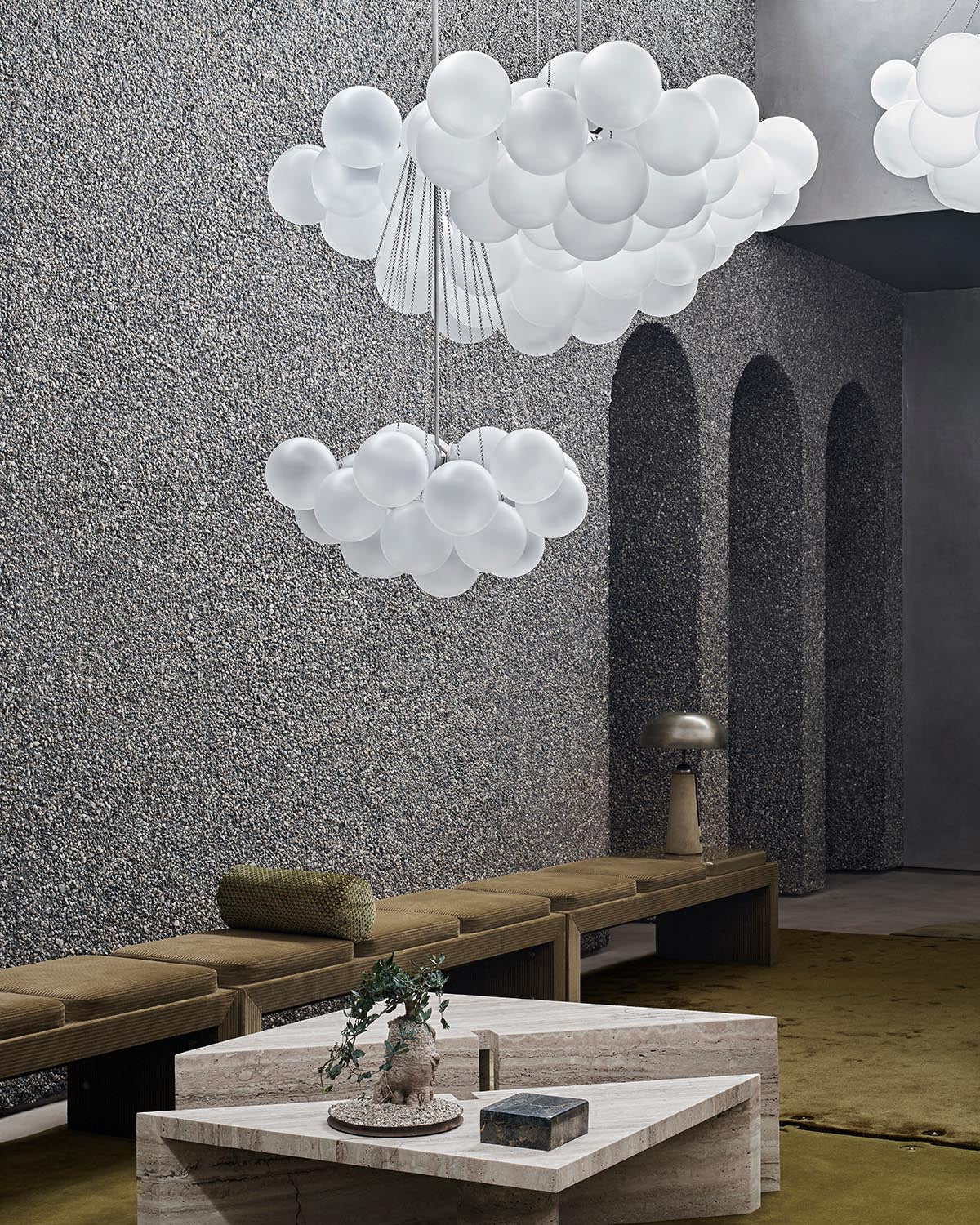 Contemporary seating area with textured gray walls, olive green benches, geometric stone tables, and cloud-like hanging lights