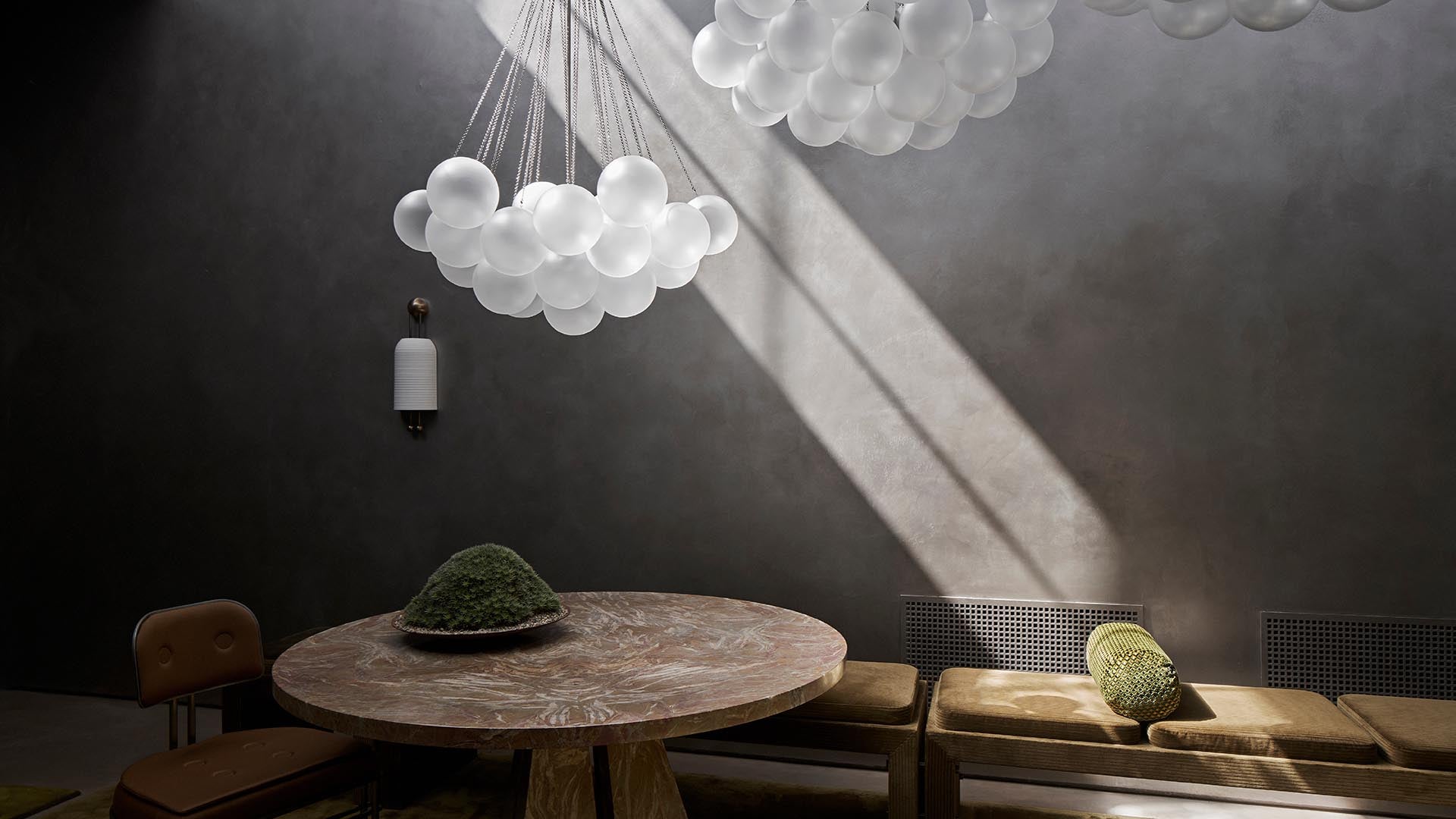 Sunlight streaming through a window illuminates a round wooden table with moss centerpiece and surrounding brown chairs on gray walls