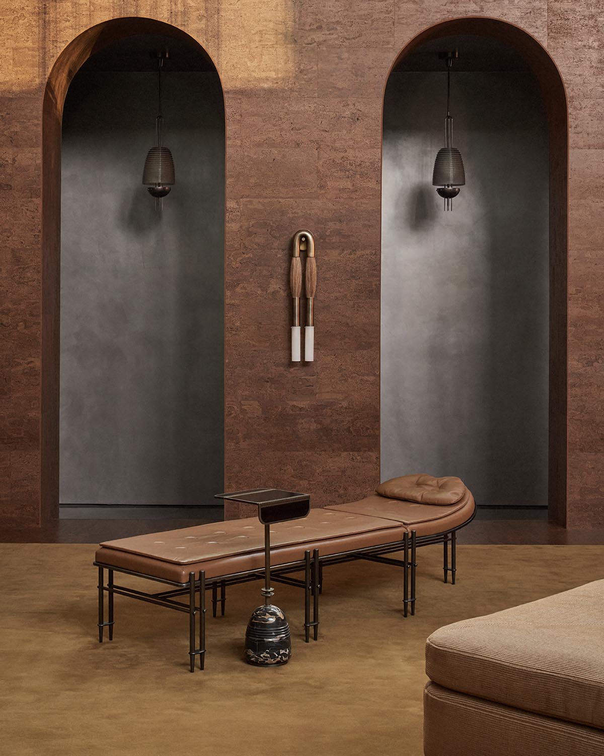 Curved brown leather bench with cylindrical legs set against a textured brown wall with two arched doorways and hanging lights